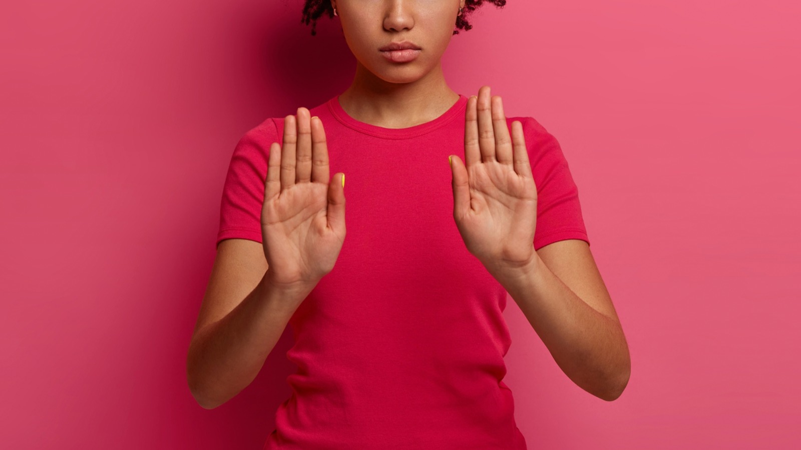 Foto ilustrativa colorida mostra uma mulher nera usando uma blusa rosa fazendo o sinal de "pare" com as duas mãos.