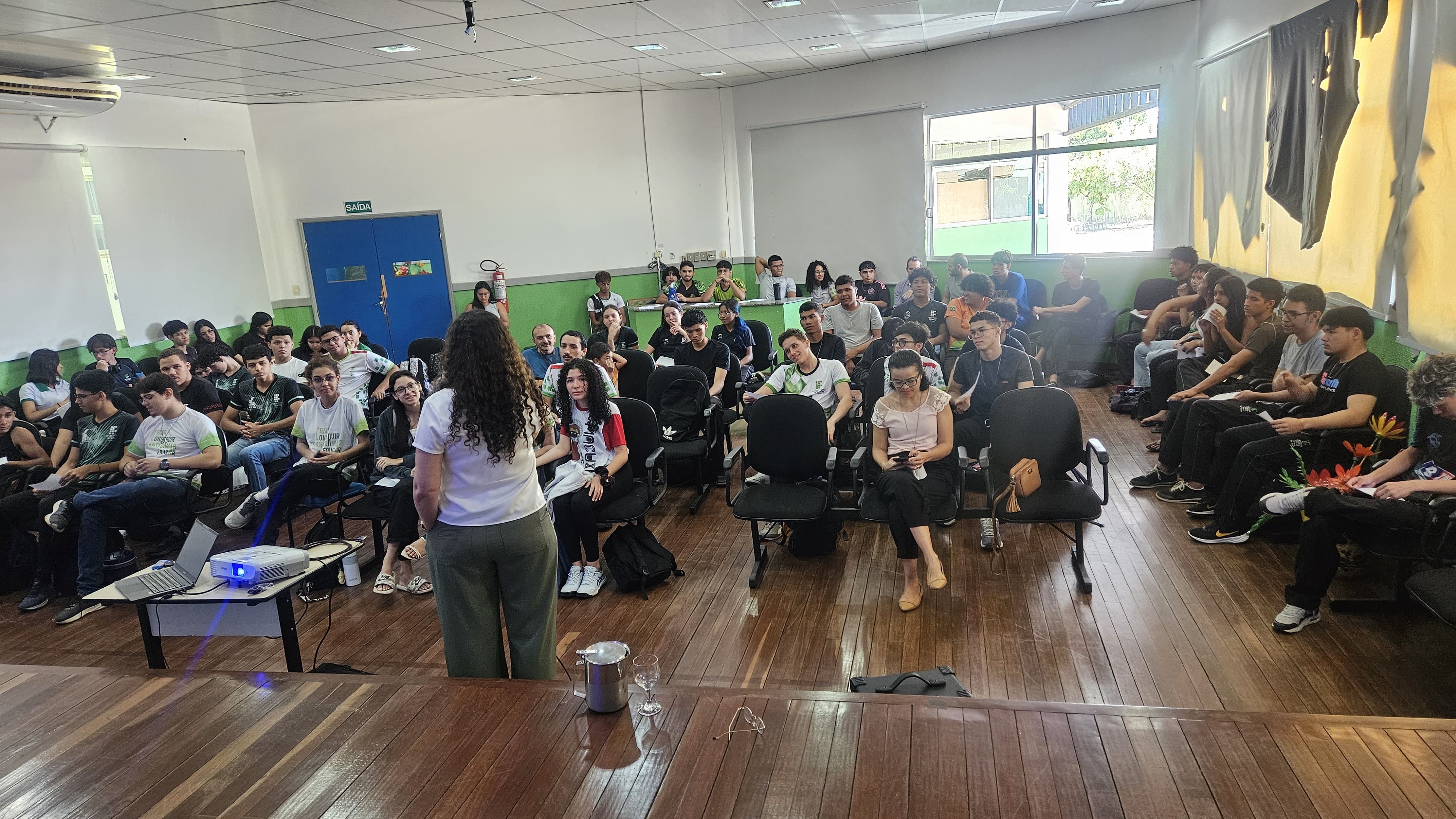 Imagem colorida contém a coordenadora do programa Justiça Comunitária do TJRR, Marcelle Grécia Wottrich, palestrando para dezenas de adolescentes em uma sala de aula. Abaixo a frase “PROMOÇÃO DA PAZ SOCIAL - Justiça Comunitária do TJRR encerra 2024 com resultados expressivos no 4º trimestre.”