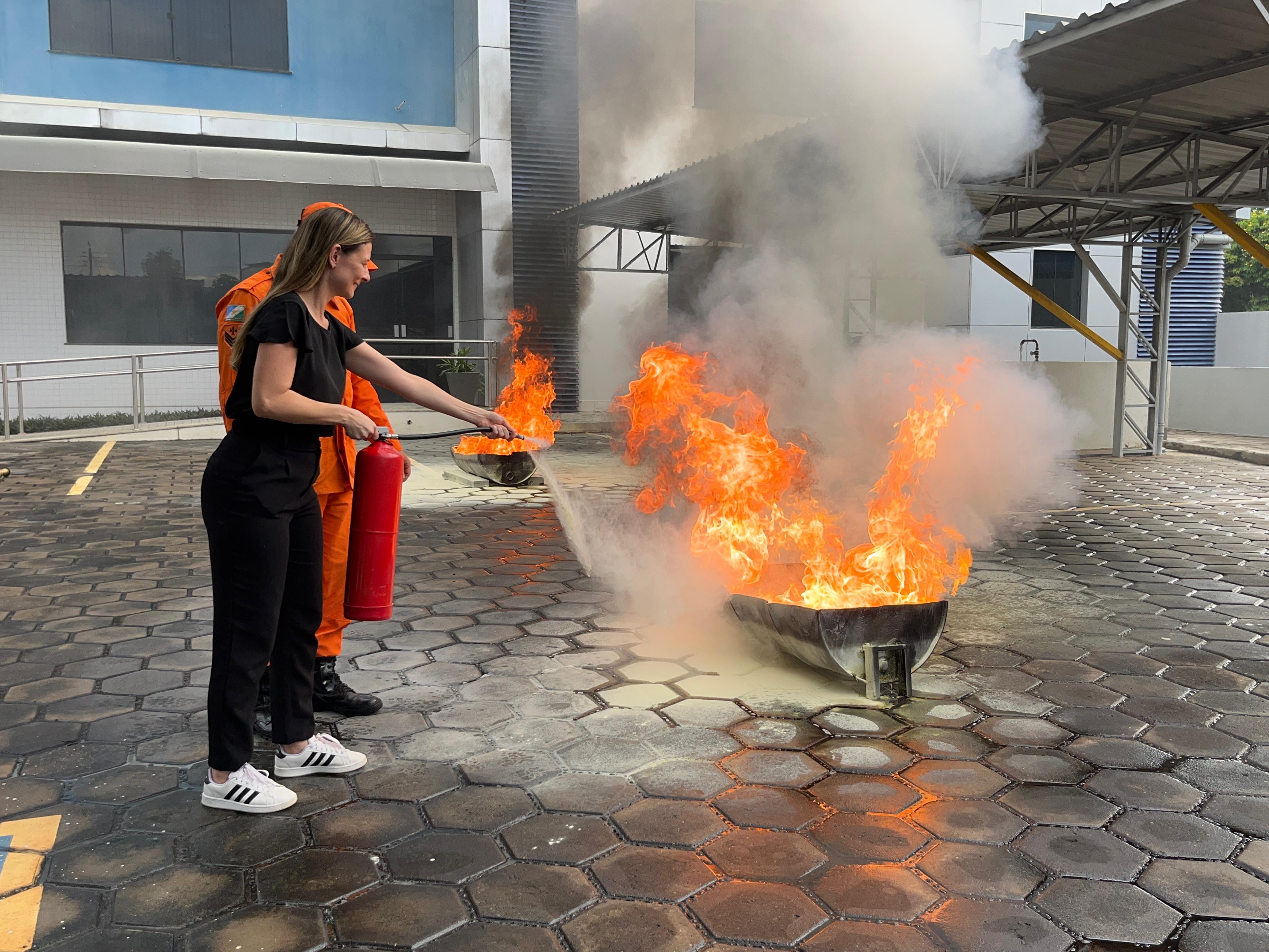 Foto colorida mostra um bombeiro auxiliando uma servidora a usar o extintor de incêndio durante o treinamento de brigadista