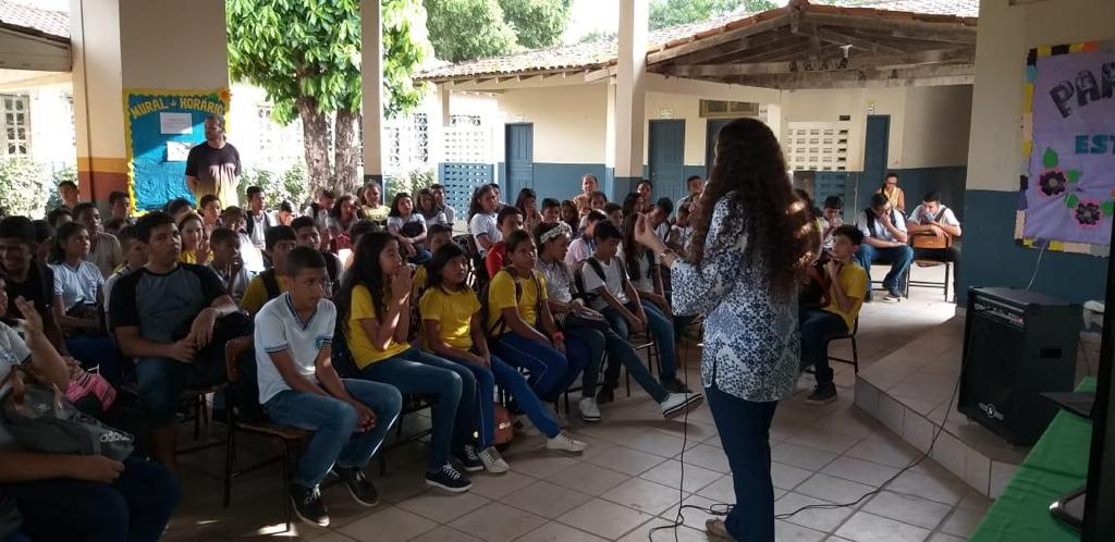 Mulher de pé falando no microfone ligado à caixa de som em pátio de escola, com vários alunos sentados em cadeiras olhando e ouvindo ela.