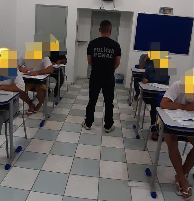 Foto de detentos sentados fazendo uma prova, com um policial penal em pé de costas, no meio da sala.