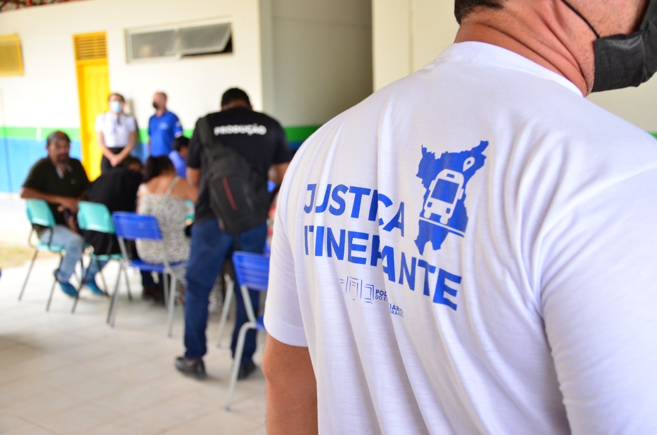 Imagem colorida contém homem de costas usando máscara com a frase na camisa “justiça itinerante”, com fundo da imagem da imagem embasado contendo pessoas
