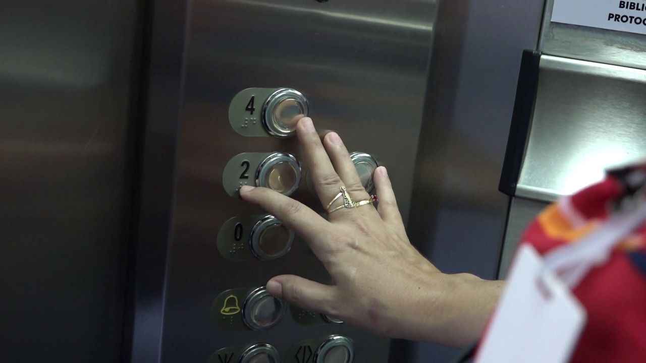 Imagem colorida mostra mão apertando botões de elevador contendo sinais de braille nos botões. 