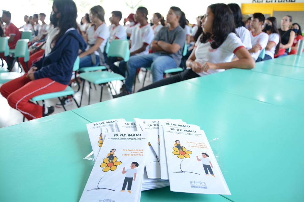 Imagem colorida contém alunos sentados enfileirados durante palestra de conscientização da equipe da Divisão de Proteção das Varas da Infância e Juventude do TJRR, com flyers da ação em uma mesa ao lado dos alunos. 