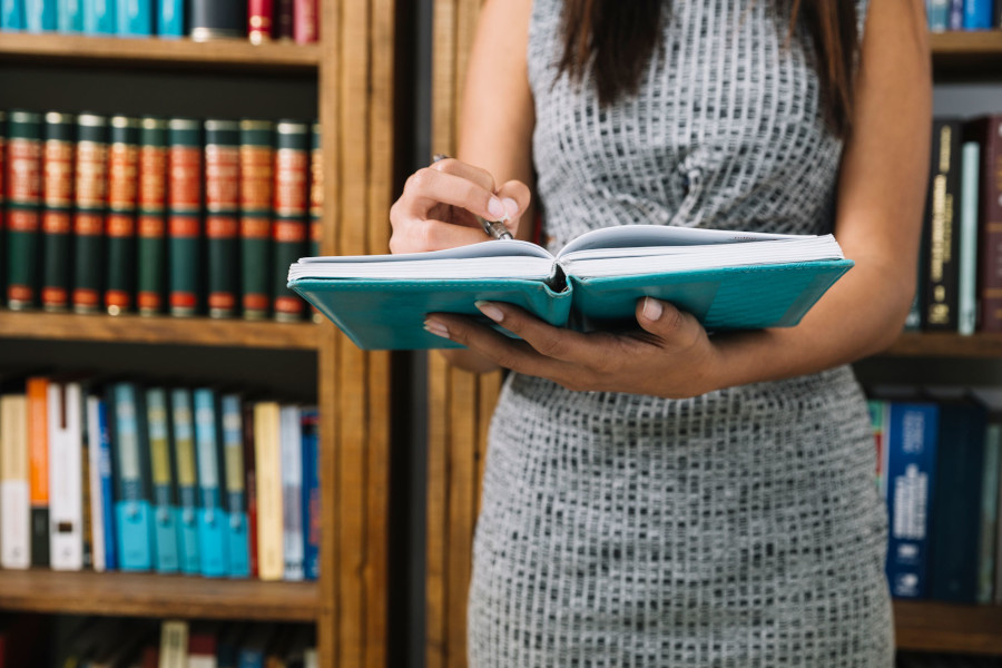 Imagem colorida contém uma mulher assinando um caderno. Atrás dela contém uma estante contendo livros. 