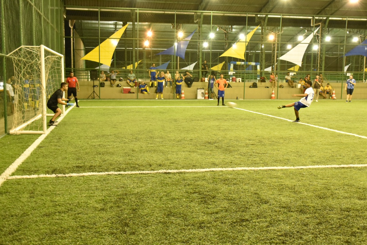 Imagem colorida contém jogador de futsal chutando a bola na direção da rede e o goleiro em posição de defesa.