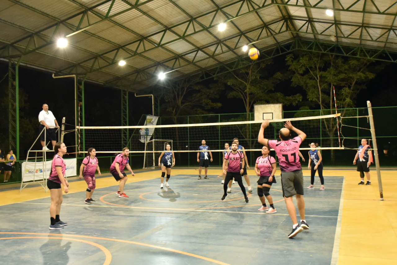 Imagem colorida contém dois times de vôlei, disputando partida na quadra.