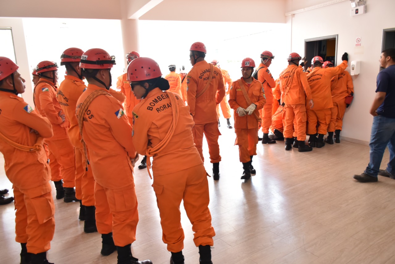 Imagem colorida mostra 26 alunos  Cabos  com capacetes e  roupas no tom laranja em pé em uma sala.