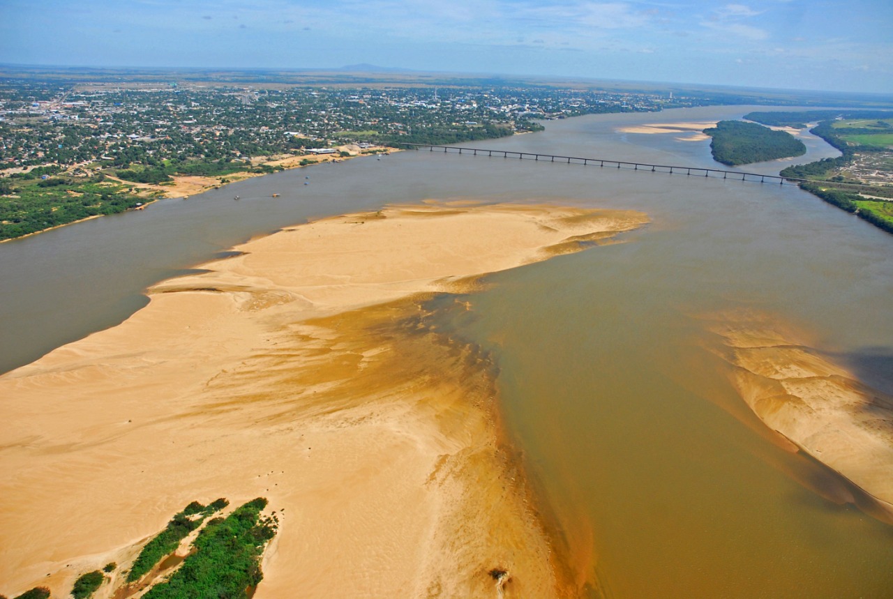 Imagem colorida contém a vista do drone de cima  do Rio Branco com bancos de areia e ao fundo da imagem contém a Ponte dos Macuxis. 