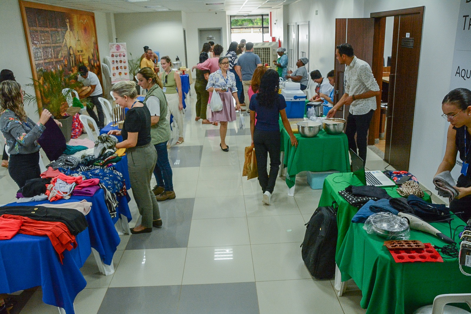 foto colorida mostra a feira de orgânicos e bazar do desapego no hall do Fórum Criminal. Os vendedores e expositores estão com mesas montadas dos lados direito e esquerdo do hall. Ao meio transitam os compradores da feira. É possível observar mesas com vendas de roupas, utensílios de decoração, comida e artesanato indígena.