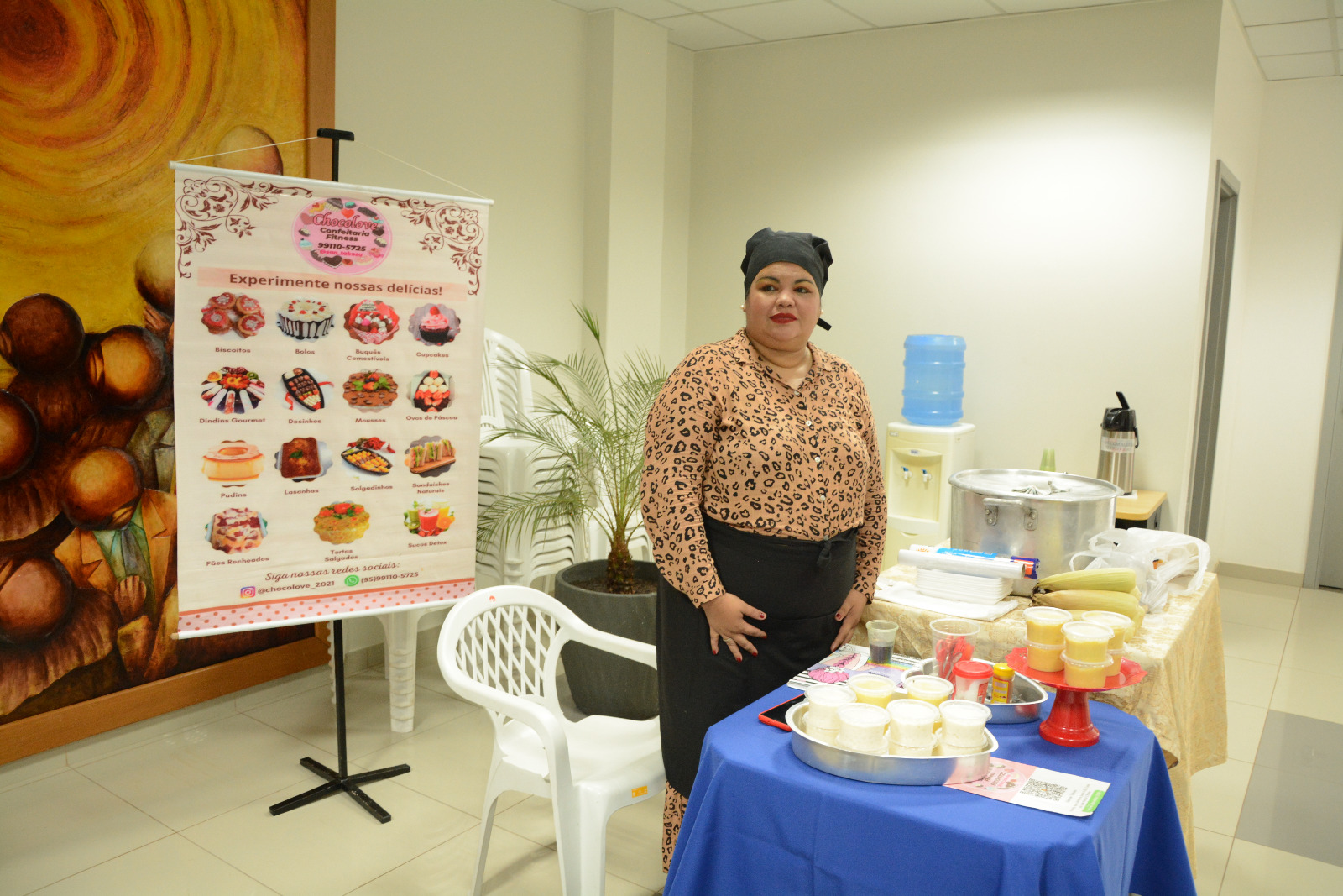 foto colorida mostra uma mulher em pé, em frente a mesa onde estão expostos seus produtos como canjica, milho e mungunzá. Atrás dela está um banner com os produtos vendidos pela confeitaria Chocolove. A mulher usa blusa com estampa de onça, toca e saia preta. 