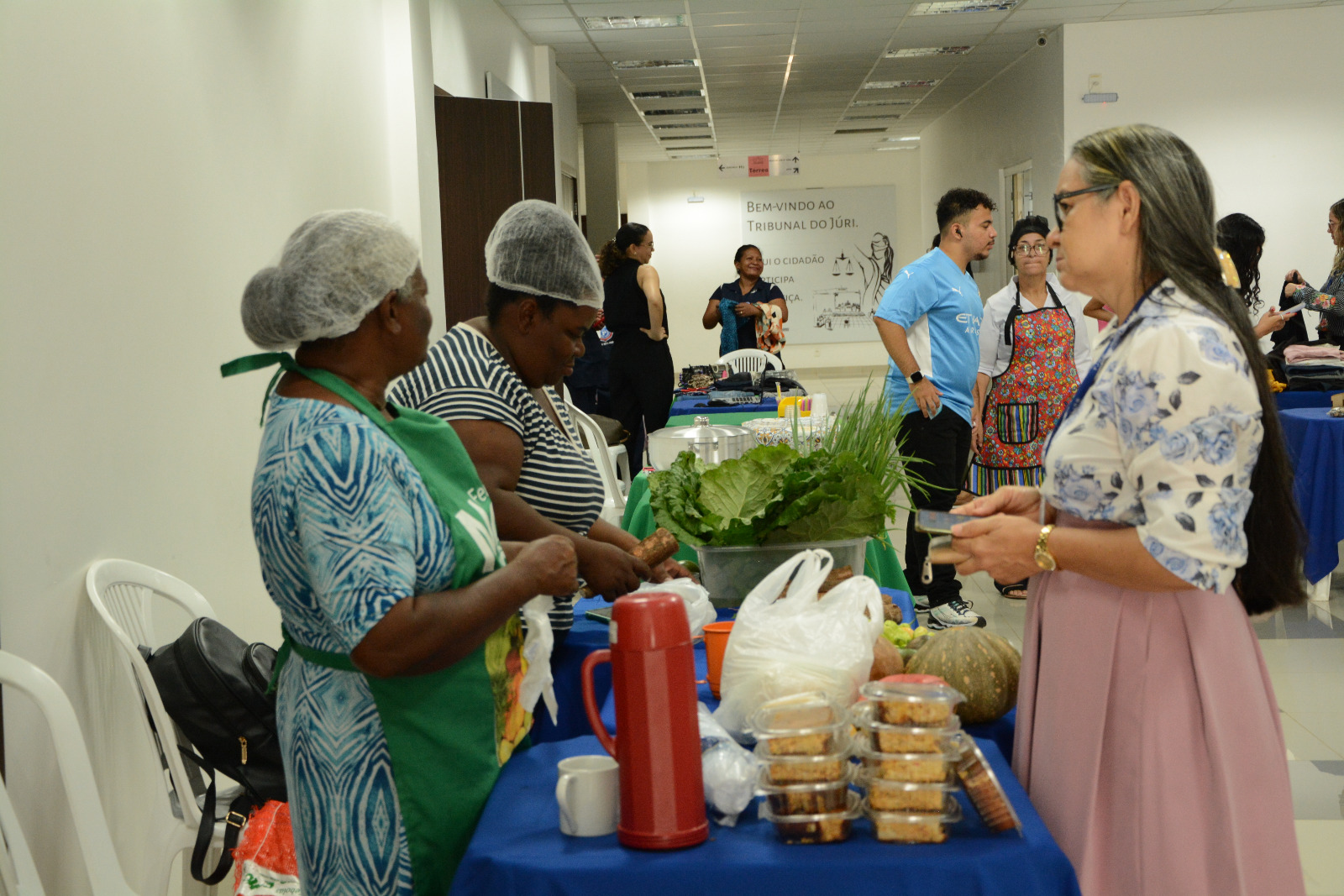  foto colorida mostra a esquerda duas mulheres produtoras rurais em pé, vendendo macaxeira para uma servidora do TJRR. Frente às duas mulheres está uma mesa com vários produtos como bolos, verduras, frutas e vegetais. 