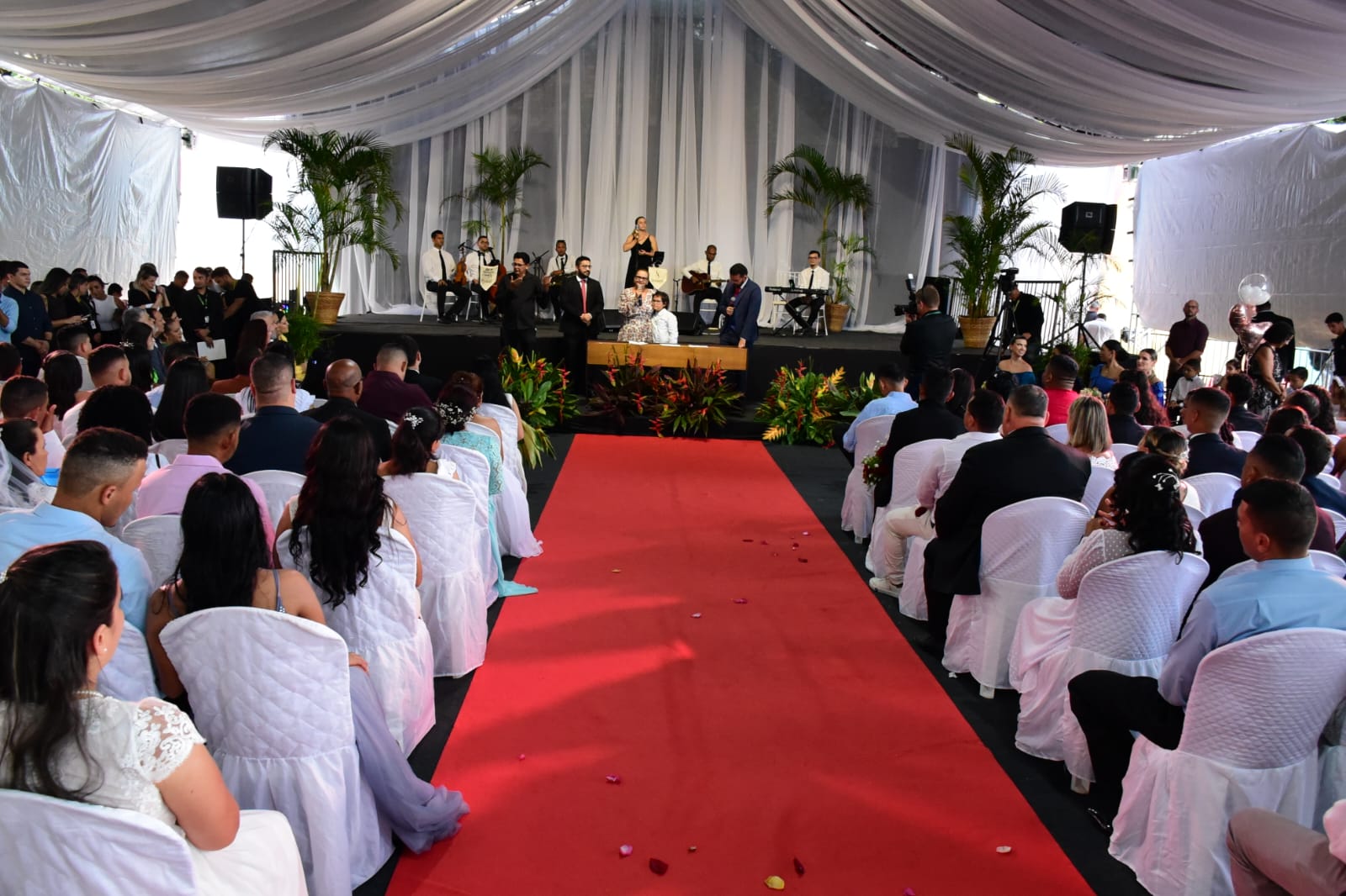  foto colorida mostra o palco da tenda onde está sendo realizada a cerimônia. Frente a ele está a juíza Graciete Sotto Mayor, posicionada em pé, atrás de uma mesa, falando ao microfone. Frente a ela está o público da plateia e um tapete vermelho.