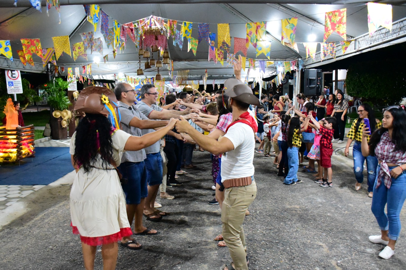 Mostra servidores dançando quadrilha em meio a um local decorado em tema de festas juninas. Eles estão de mãos dadas formando um túnel com os braços.