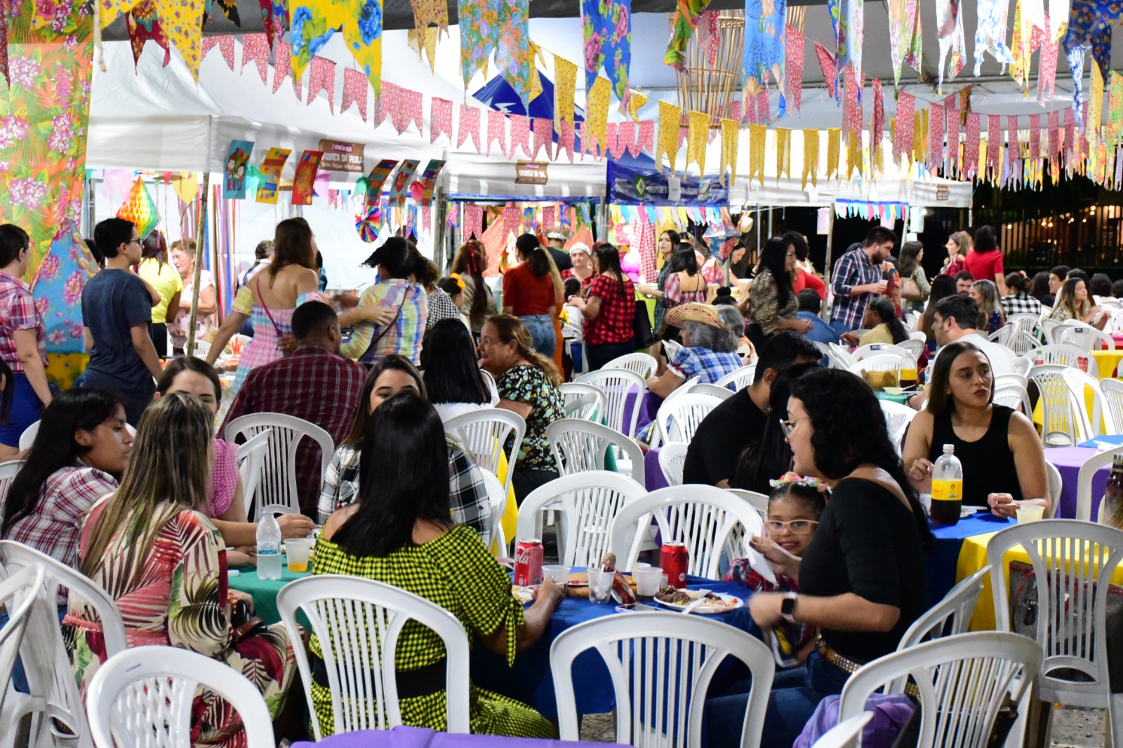Mostra dezenas de servidores e familiares sentados em uma praça de alimentação. Atrás deles, aparecem várias barraquinhas que comercializam comidas típicas.