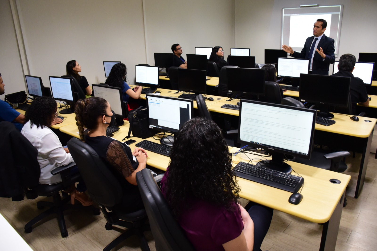 Imagem colorida mostra 11 servidores sentados em frente a computadores distribuídos em quatro fileiras. Eles observam atentos o condutor do treinamento, que está em pé à frente deles, trajando terno e gravata.