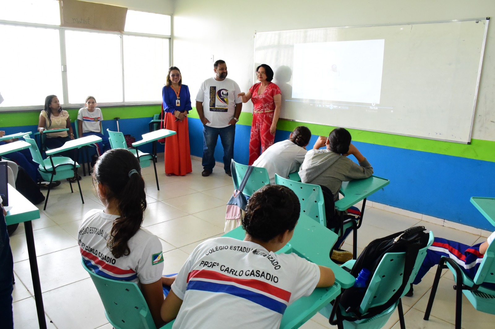 foto colorida em uma sala de aula com 3 pessoas falando para a turma de alunos