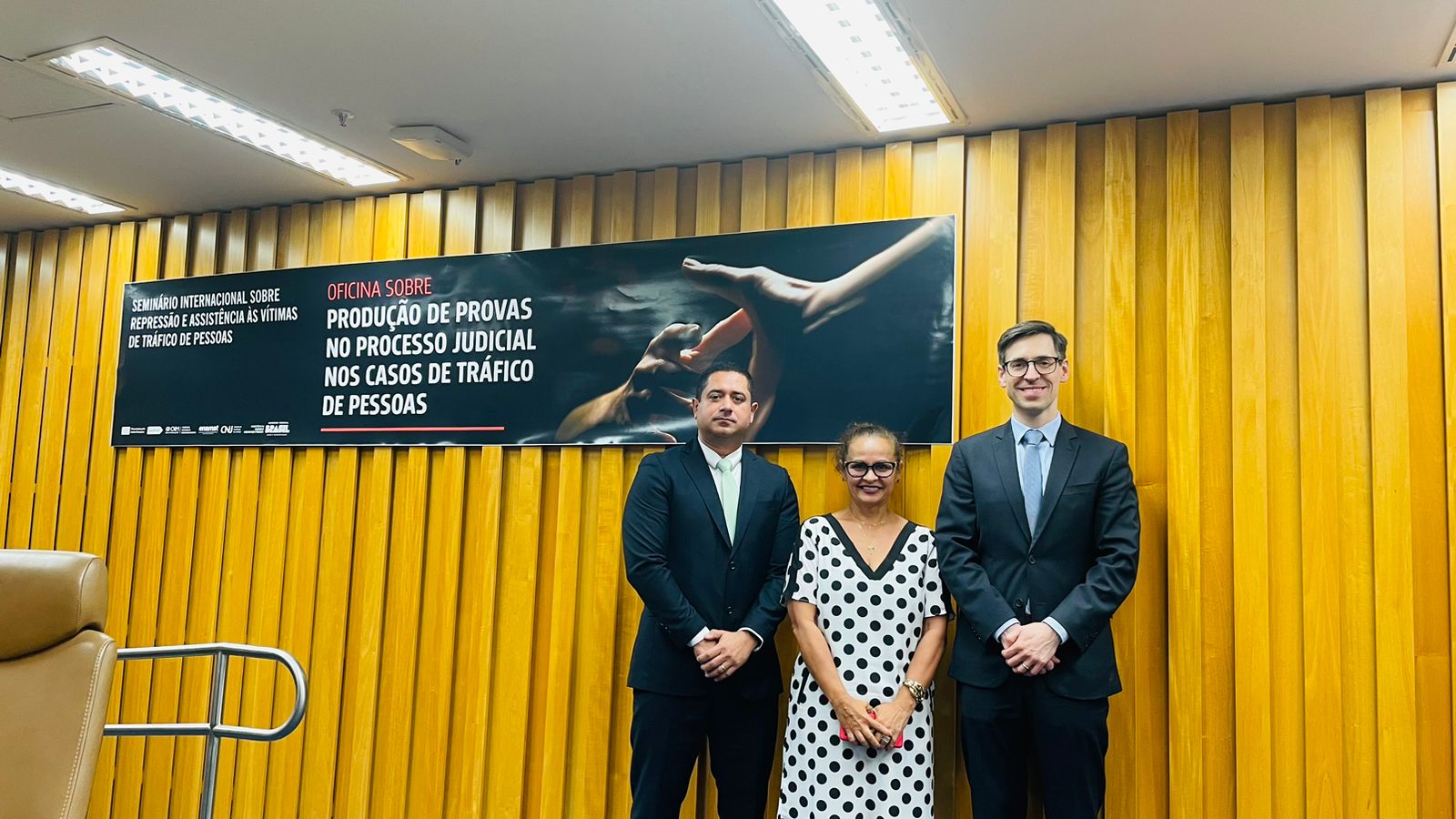 Foto colorida da Juíza Graciete Sotto Mayor Ribeiro, ao lado de dois homens de terno e a frente do banner "oficina sobre produção de provas no processo judicial nos casos de trafico de pessoas"