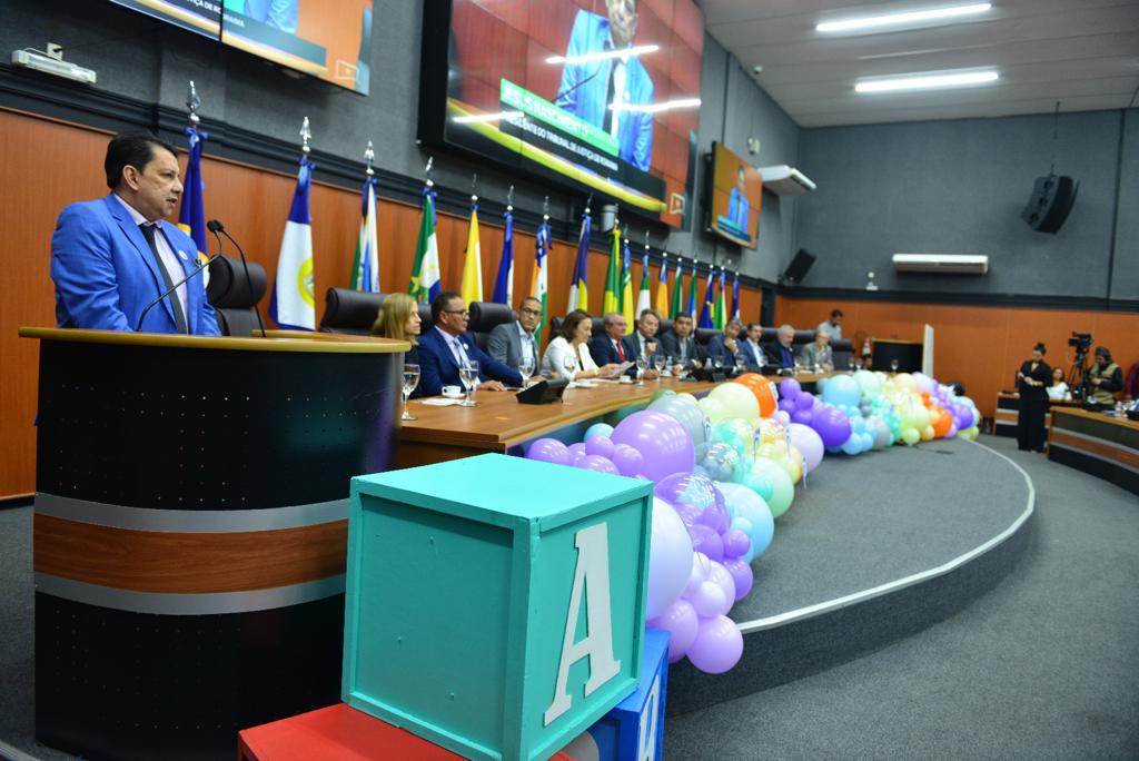  foto colorida mostrando o Presidente do TJRR, desembargador Jésus Nascimento no pulpito, ao lado está a mesa de autoridades compostas por representantes do Legislativo, Executivo e Judiciário. A frente da mesa contém  balões roxos, azul, amarelo e laranja em tons claros. 