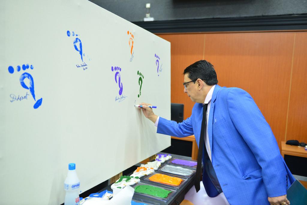  foto colorida mostrando o Presidente do TJRR, desembargador Jésus Nascimento, assinando uma lousa branca com um pincel azul. Na lousa possui algumas assinaturas seguidas de carimbos coloridos em formato de pé. 