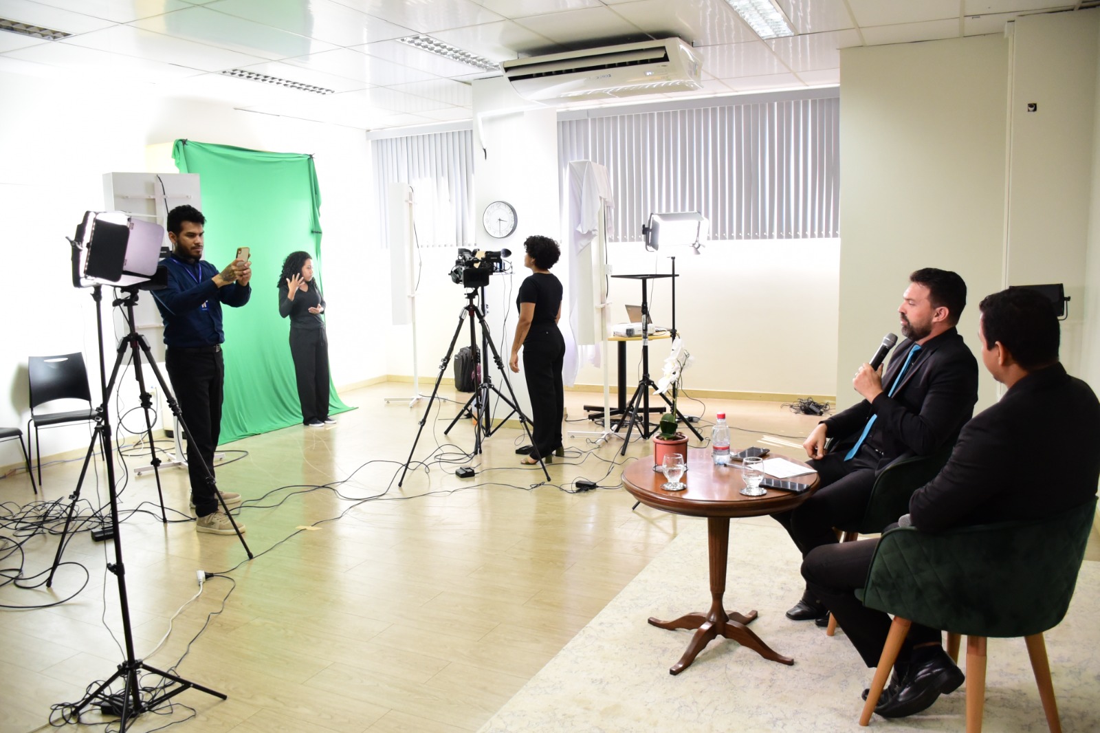  Foto colorida do ambiente onde ocorre a filmagem dos intérpretes. Na imagem contém dois intérpretes sentados e mais ao fundo possui um pano verde na qual há uma intérprete mulher gravando e outra na câmera.