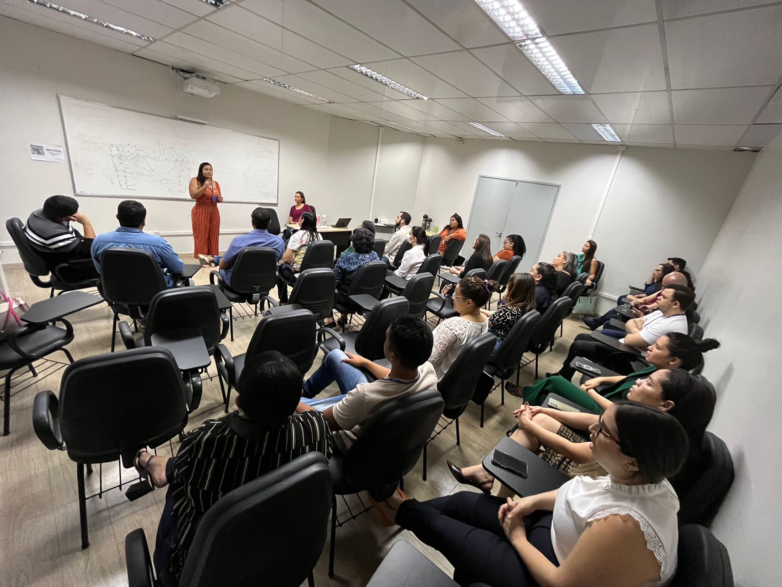  Imagem colorida  mostra a psicóloga do SQV/TJRR, Perla Lima e a Secretária do SQV/TJRR, Ivy Amaro palestrando em pé, para um grupo de pessoas sentadas em cadeiras de frente para ela, em uma sala.