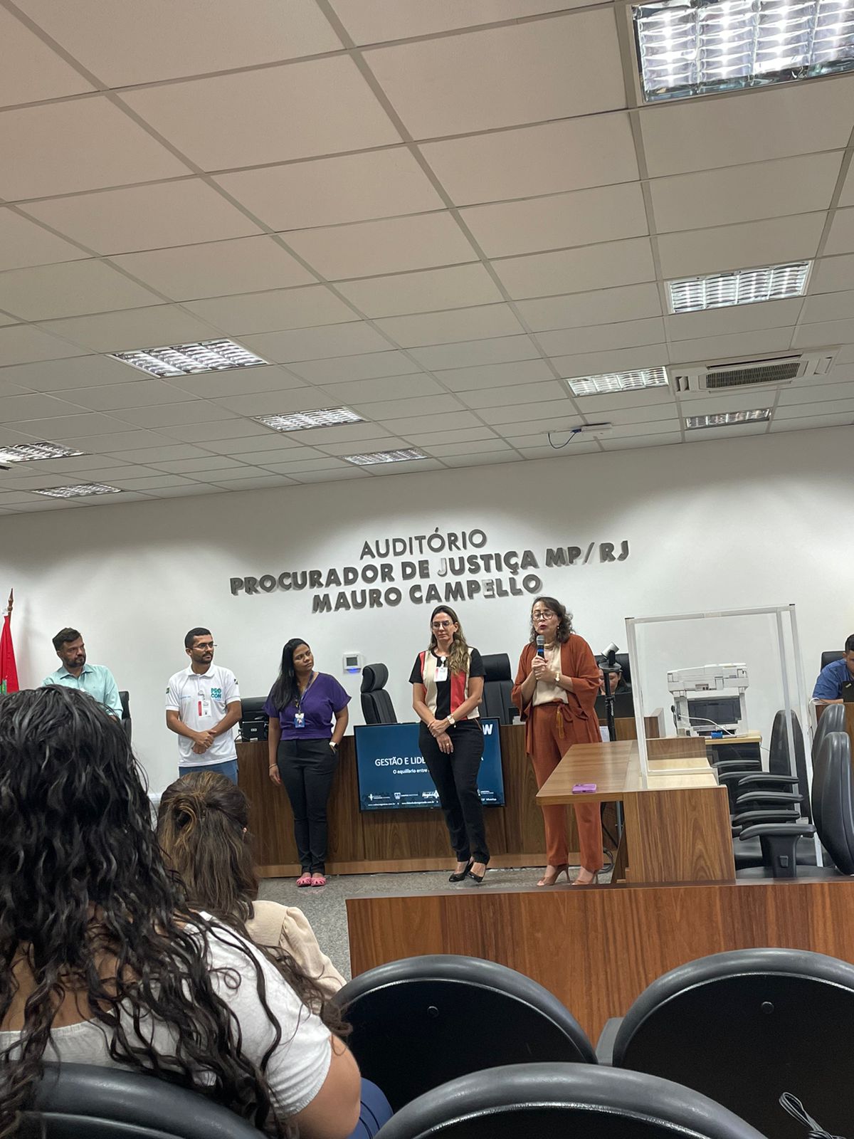 foto colorida mostra a diretora do Fórum Criminal Ministro Evandro Lins e Silva, juíza Lana Leitão, ao lado da reitora do Centro Universitário Estácio da Amazônia, Brena Linhares, da psicóloga do TJRR, Perla Lima, do Secretário de Comunicação do Estado de Roraima, Weber Negreiros, do tradutor de libras e do servidor do Procon, durante abertura da  “Ação Cuidando do Servidor”, que ocorreu no  no Auditório Procurador de Justiça MP/RJ Mauro Campello, do Fórum Criminal Ministro Evandro Lins e Silva do TJRR, nas poltronas estão os servidores do TJRR assistindo a abertura.