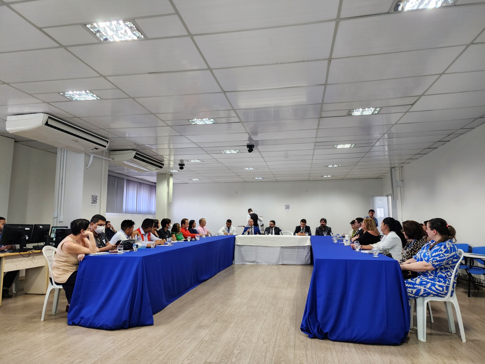 Foto colorida mostrando pessoas sentadas em frente a uma mesa de reunião. 