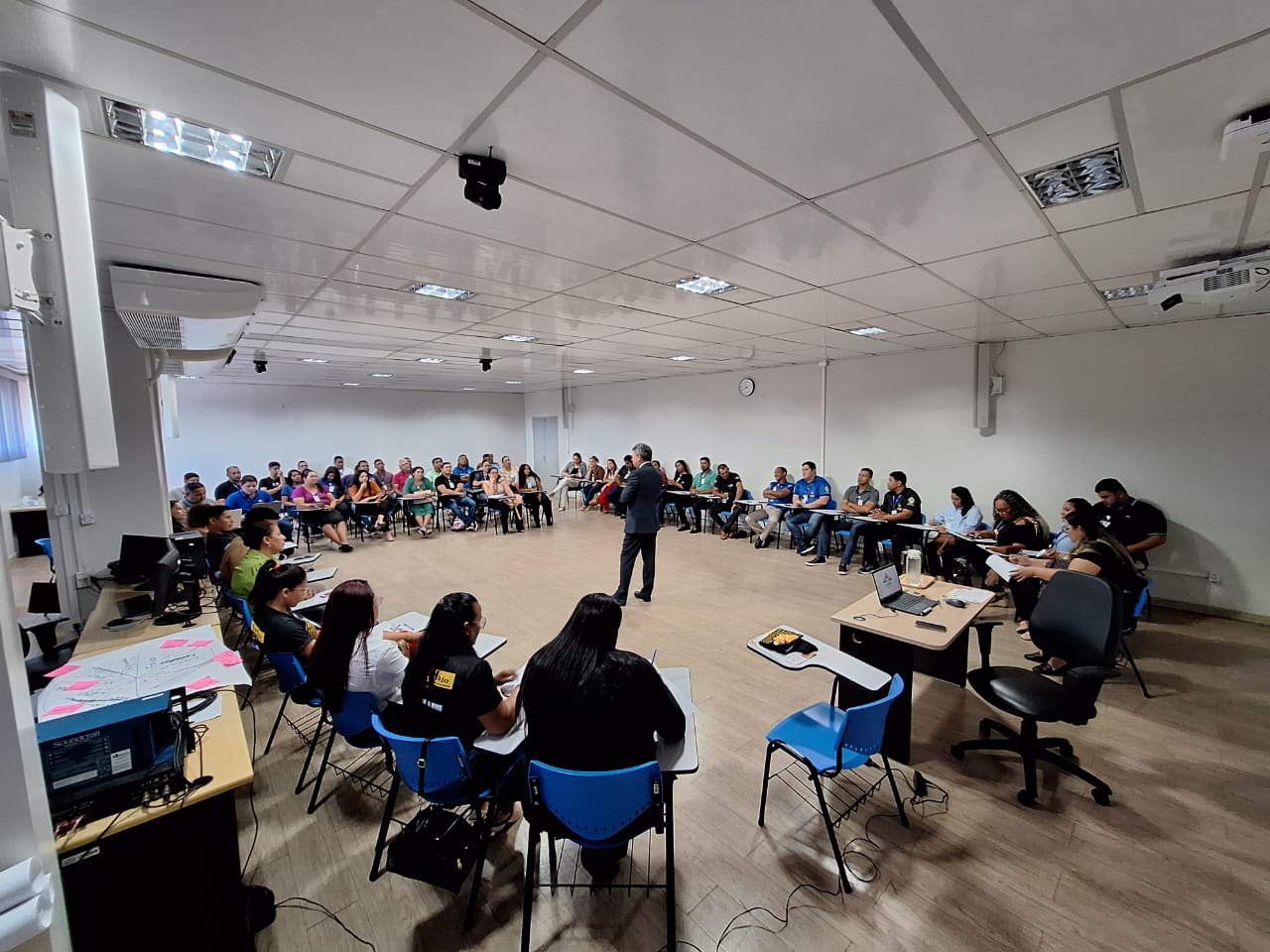 Imagem colorida de uma sala cheia de pessoas sentadas em circulo durante palestra