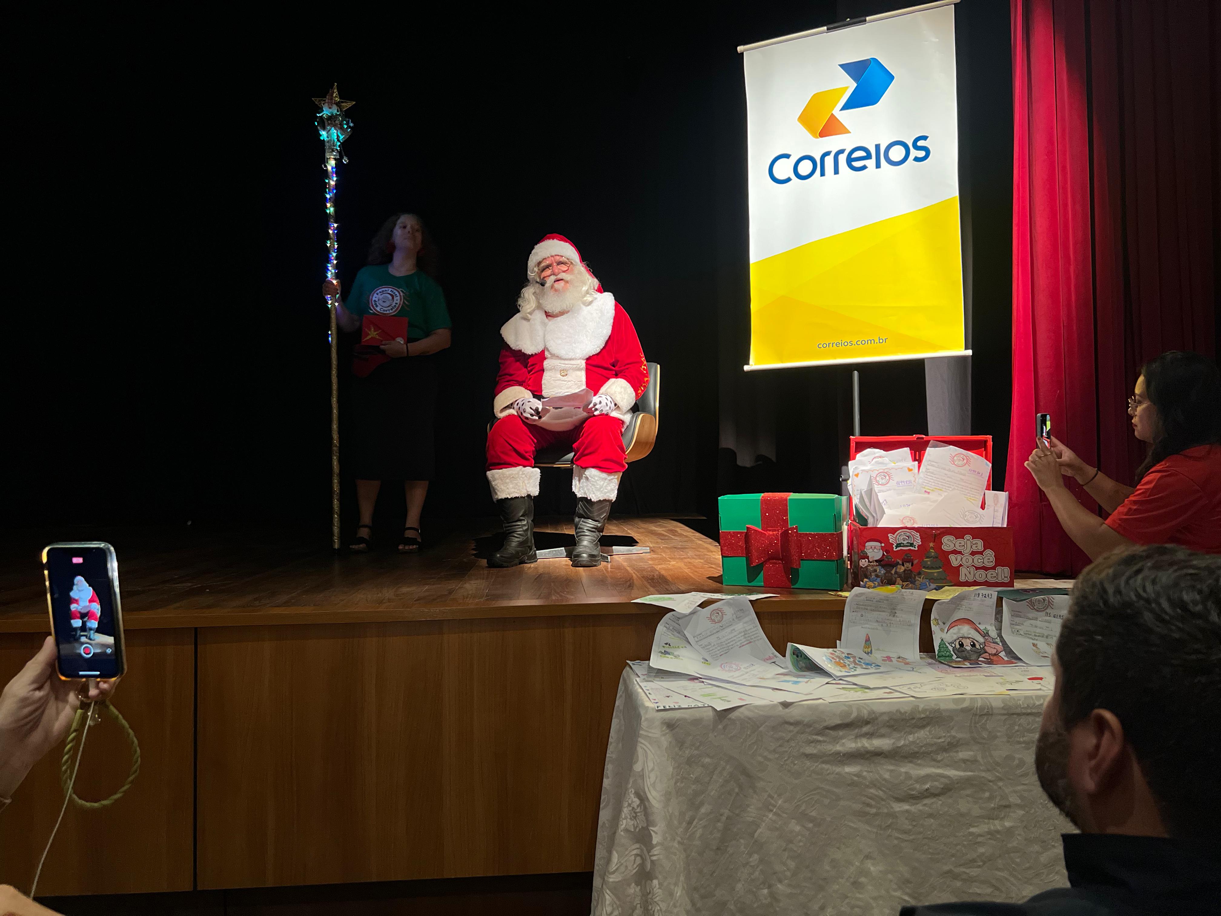 foto colorida do papai noel sentado em frente ao banner dos correios. 