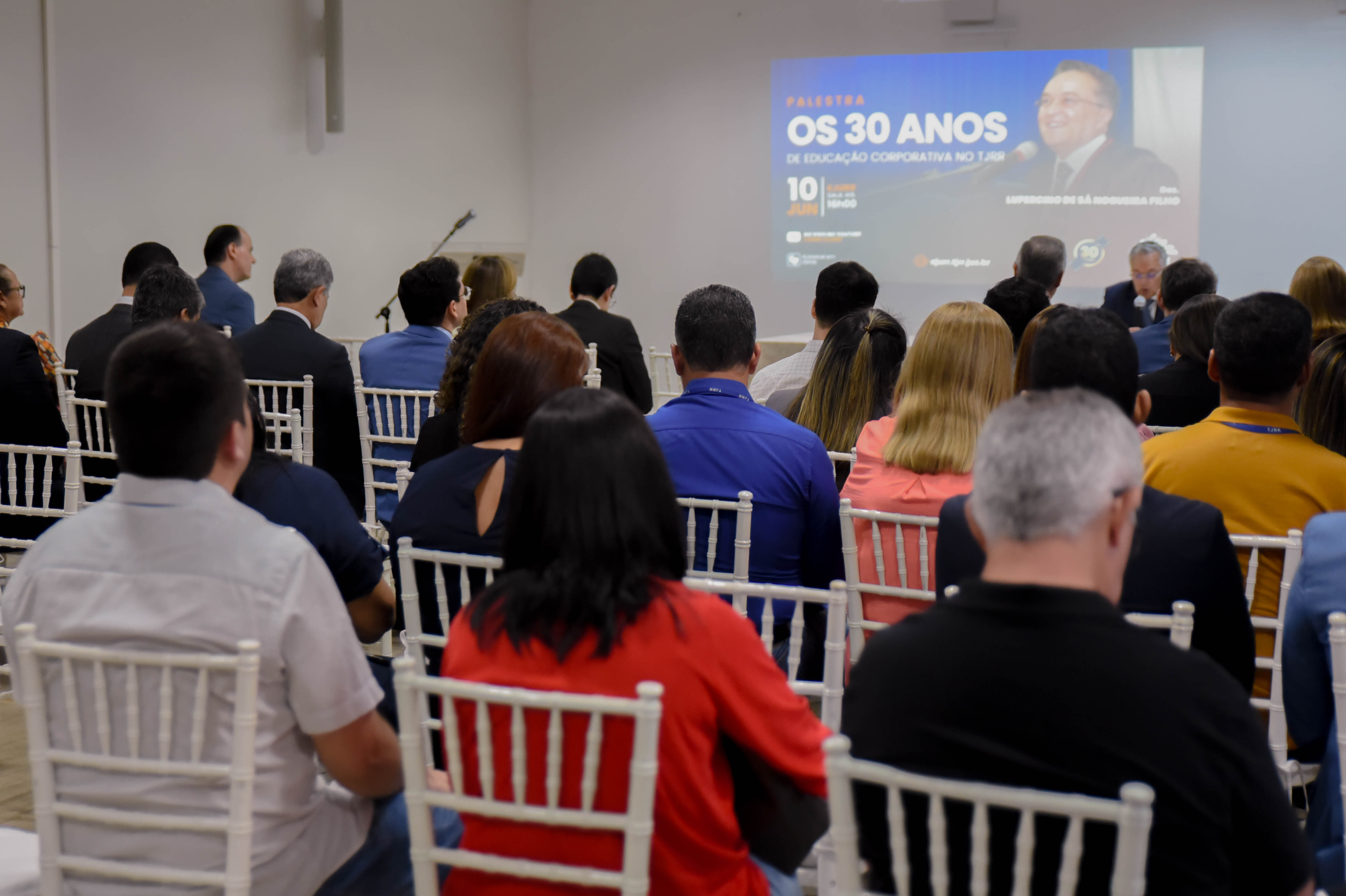 imagem colorida contém pessoas assistindo palestra todas de costas para a câmera fotografica. 