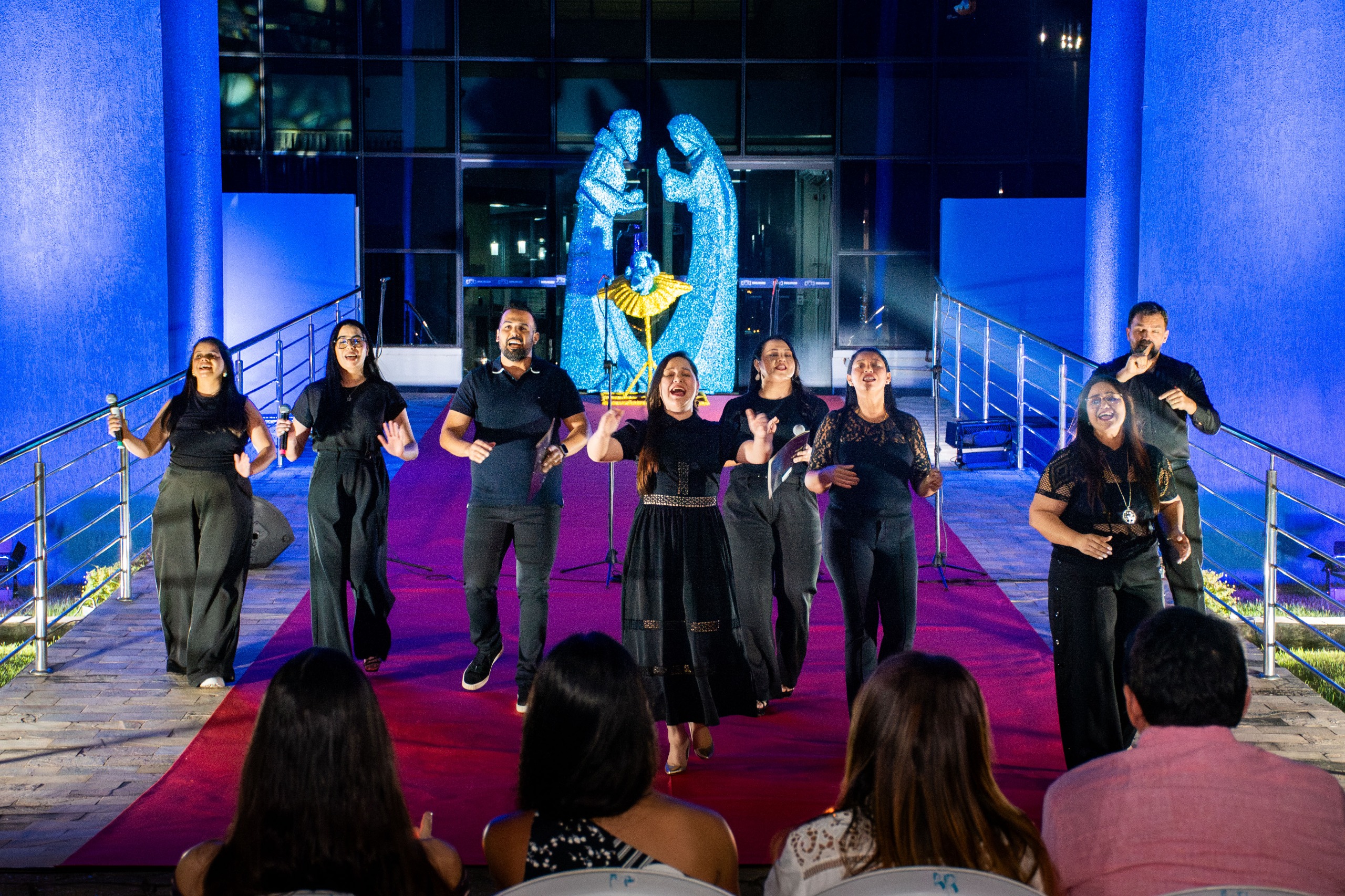 Imagem colorida mostra um coral musical contendo dez cantores se apresentando na entrada do  Palácio da Justiça Desembargador Robério Nunes dos Anjos, durante a cantata natalina do TJRR, sendo quatro homens e seis mulheres.