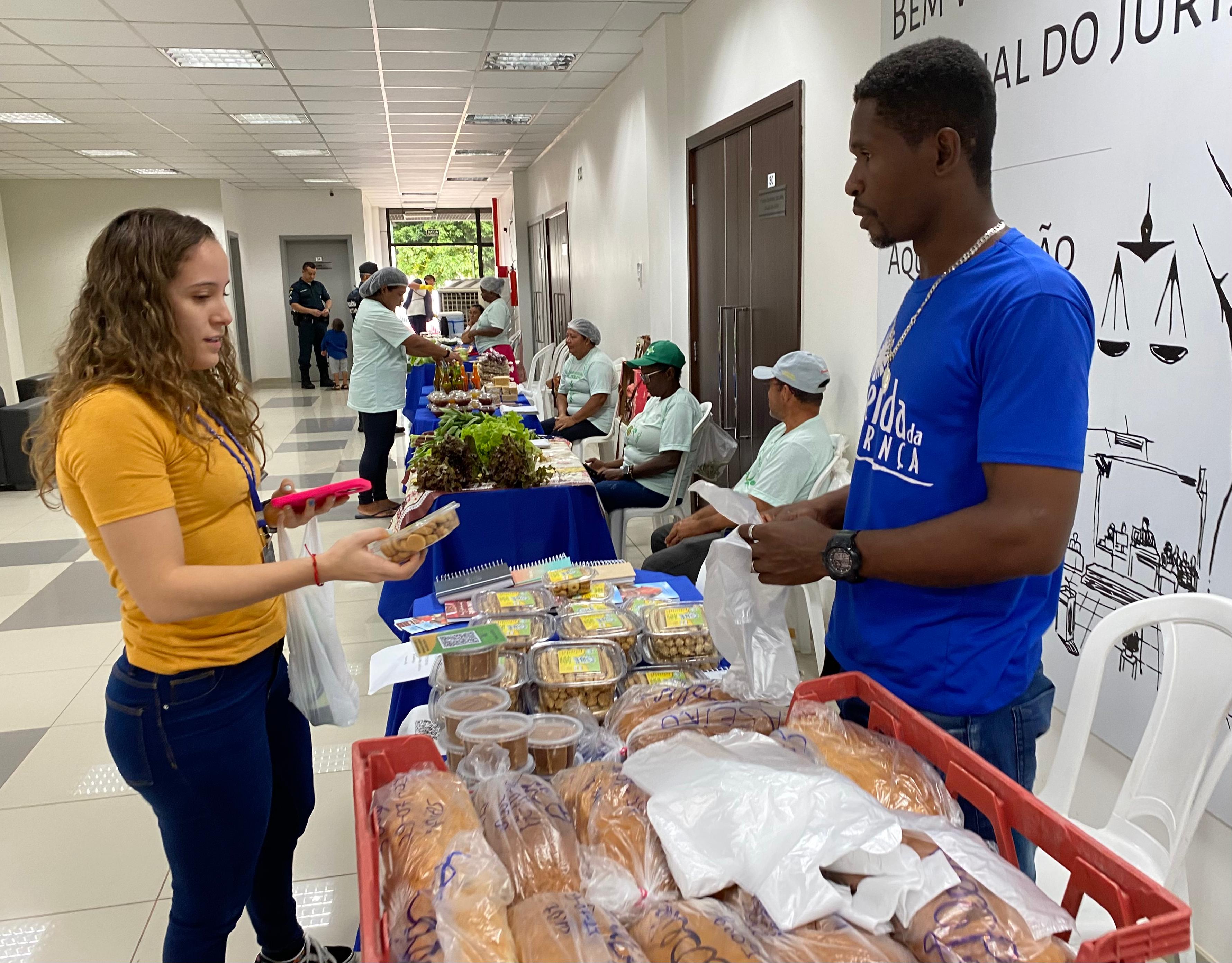 Foto colorida mostrando duas pesosas durante a venda de produtos. Uma comprando e a outra vendendo. Na imagems ainda há alimentos, em primeiro plano, como pães e doces. Em segundo plano Verduras aparecem no fundo.