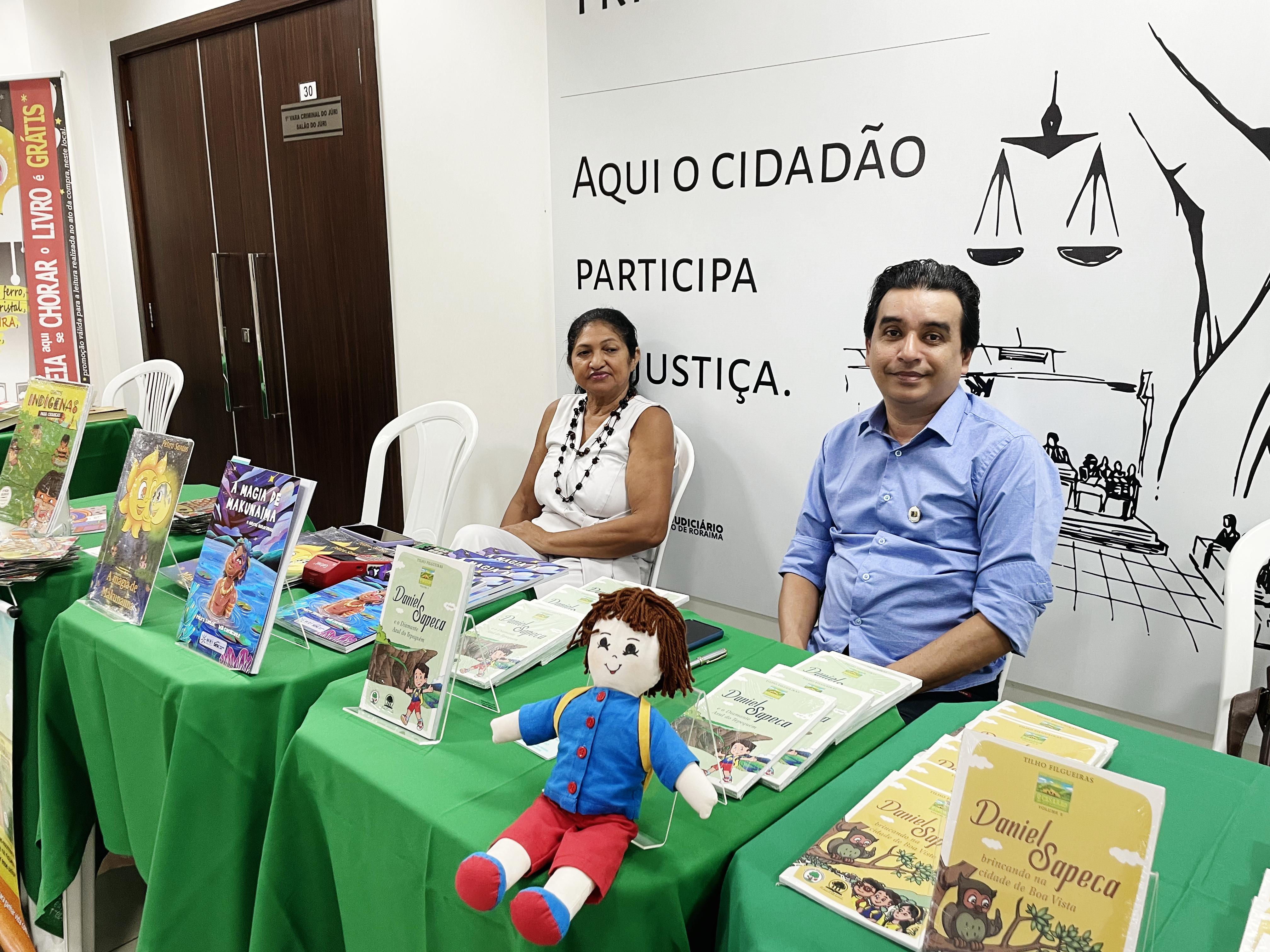 foto colorida com Clotilho Filgueiras e Pétira Santos, sentados e uma mesa à frente cheia de suas obras expostas,  posando para fotografia.