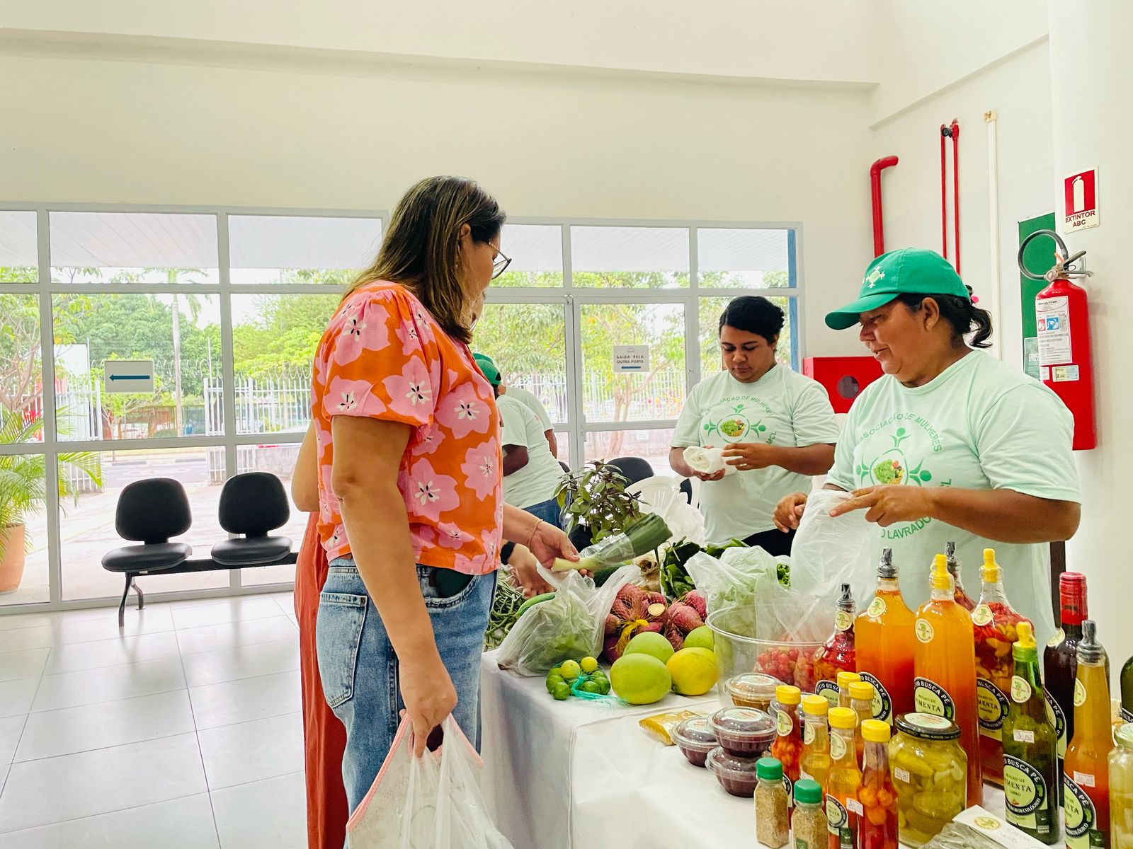 Foto colorida mostrando uma mulher vendendo alguns produtos como verduras, legumes, geleias, pimentas e entre outros. Na frente dela, duas outras mulheres estão olhando os produtos 