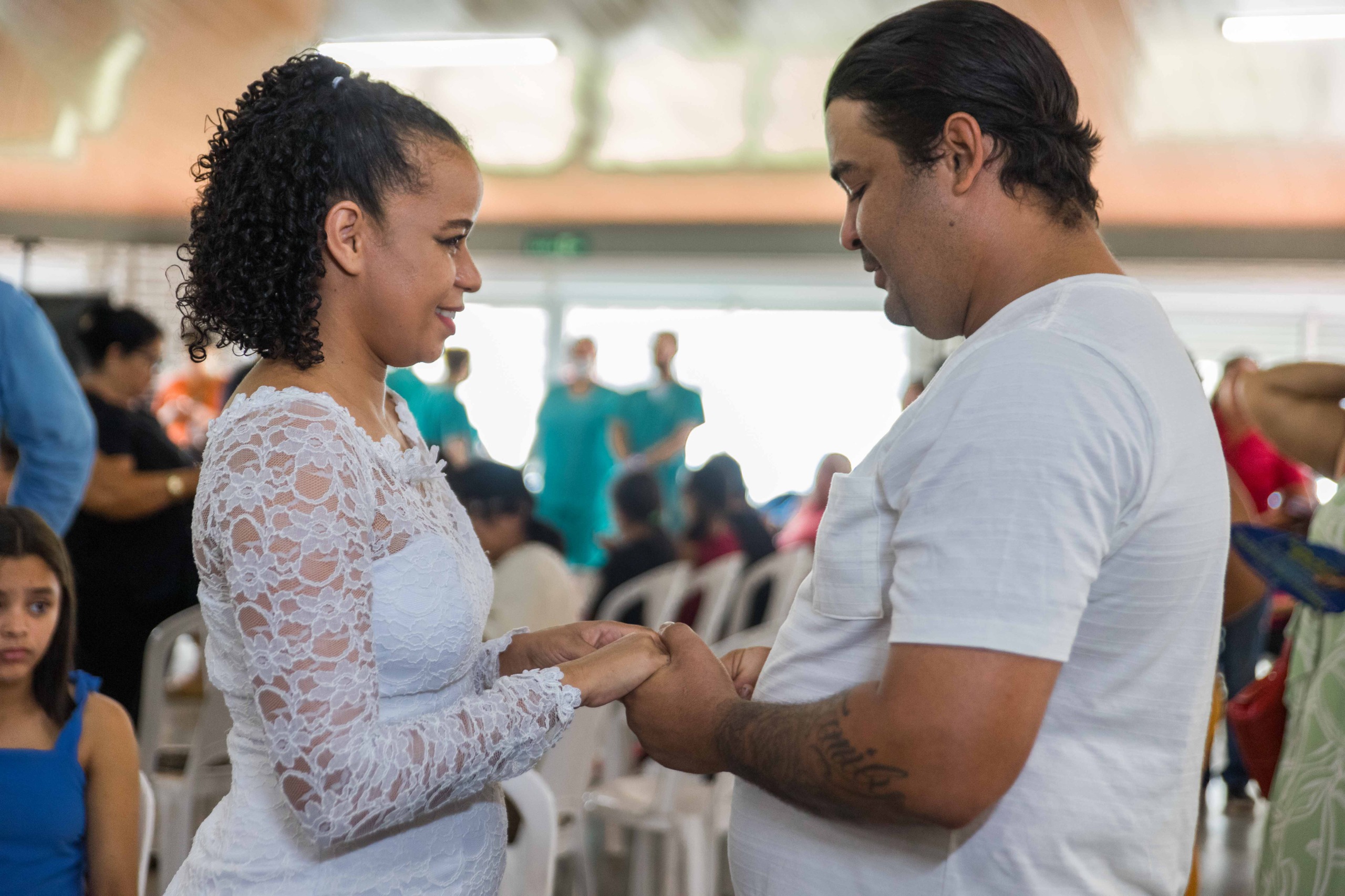 Imagem colorida mostra duas pessoas de mãos dadas durante a troca de votos na cerimônia coletiva. Ao fundo, é possível ver o público, levemente desfocado.