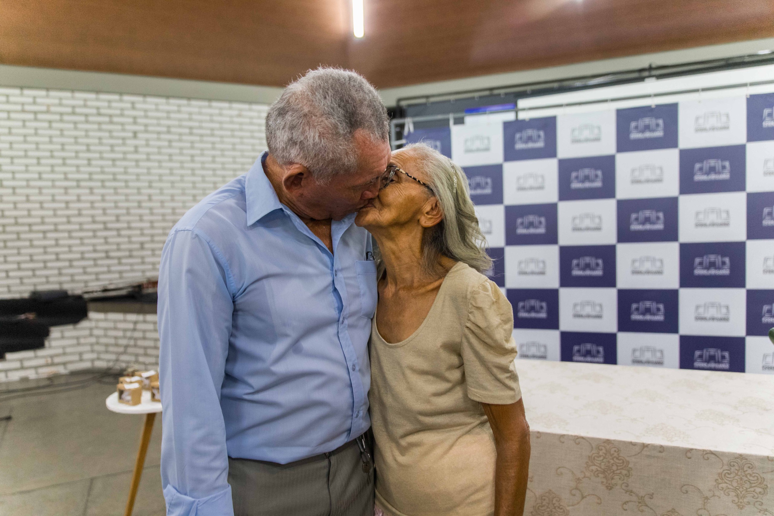 Imagem colorida mostra dois idosos se beijando durante o primeiro Casamento Coletivo promovido pela Corregedoria-Geral de Justiça de Roraima. Ao fundo, há um painel backdrop do Judiciário Roraimense e uma banqueta com bolinhos de casamento.