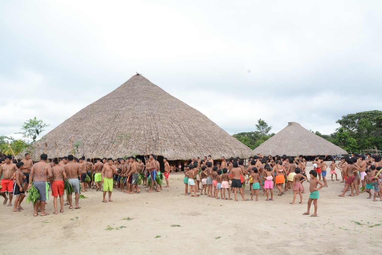 A imagem colorida mostra uma reunião de pessoas indígenas em uma área aberta, provavelmente uma aldeia. Há duas grandes construções com telhados de palha em forma cônica, características de habitações tradicionais indígenas. A imagem colorida mostra uma reunião de pessoas indígenas em uma área aberta, provavelmente uma aldeia. Há duas grandes construções com telhados de palha em forma cônica, características de habitações tradicionais indígenas.