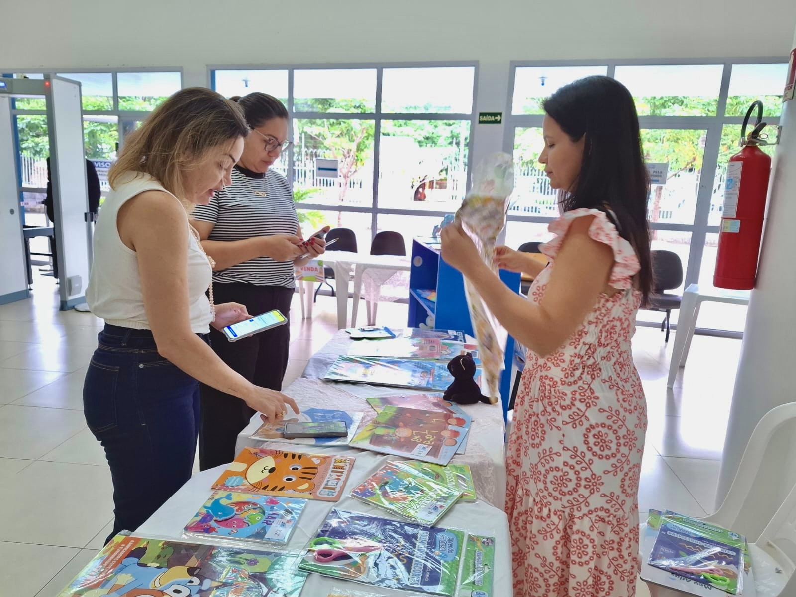 Imagem colorida contém três mulheres em vota de uma mesa com diversos livros infantis e um gato preto de pelúcia. As duas mulheres à esquerda estão utilizando o celular, realizando o pagamento via Pix. Já a mulher à direita segura um papel de presente.  Foto 3: Imagem colorida contém mesa com histórias em quadrinhos da “Turma da Mônica”. Um dos livros, à direita, aparece em destaque com o título “Mônica: a Pequena Princesa”.