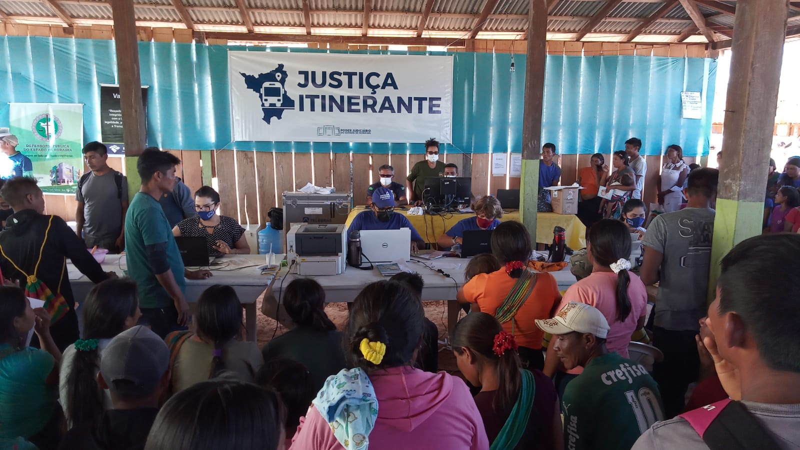 A imagem retrata uma ação da "JUSTIÇA ITINERANTE". Uma grande quantidade de pessoas, incluindo adultos e crianças, de diferentes idades e com traços indígenas visíveis, aguardam atendimento. No fundo da imagem, servidores do TJRR realizam atendimento. A imagem retrata uma ação da "JUSTIÇA ITINERANTE". Uma grande quantidade de pessoas, incluindo adultos e crianças, de diferentes idades e com traços indígenas visíveis, aguardam atendimento. No fundo da imagem, servidores do TJRR realizam atendimento.