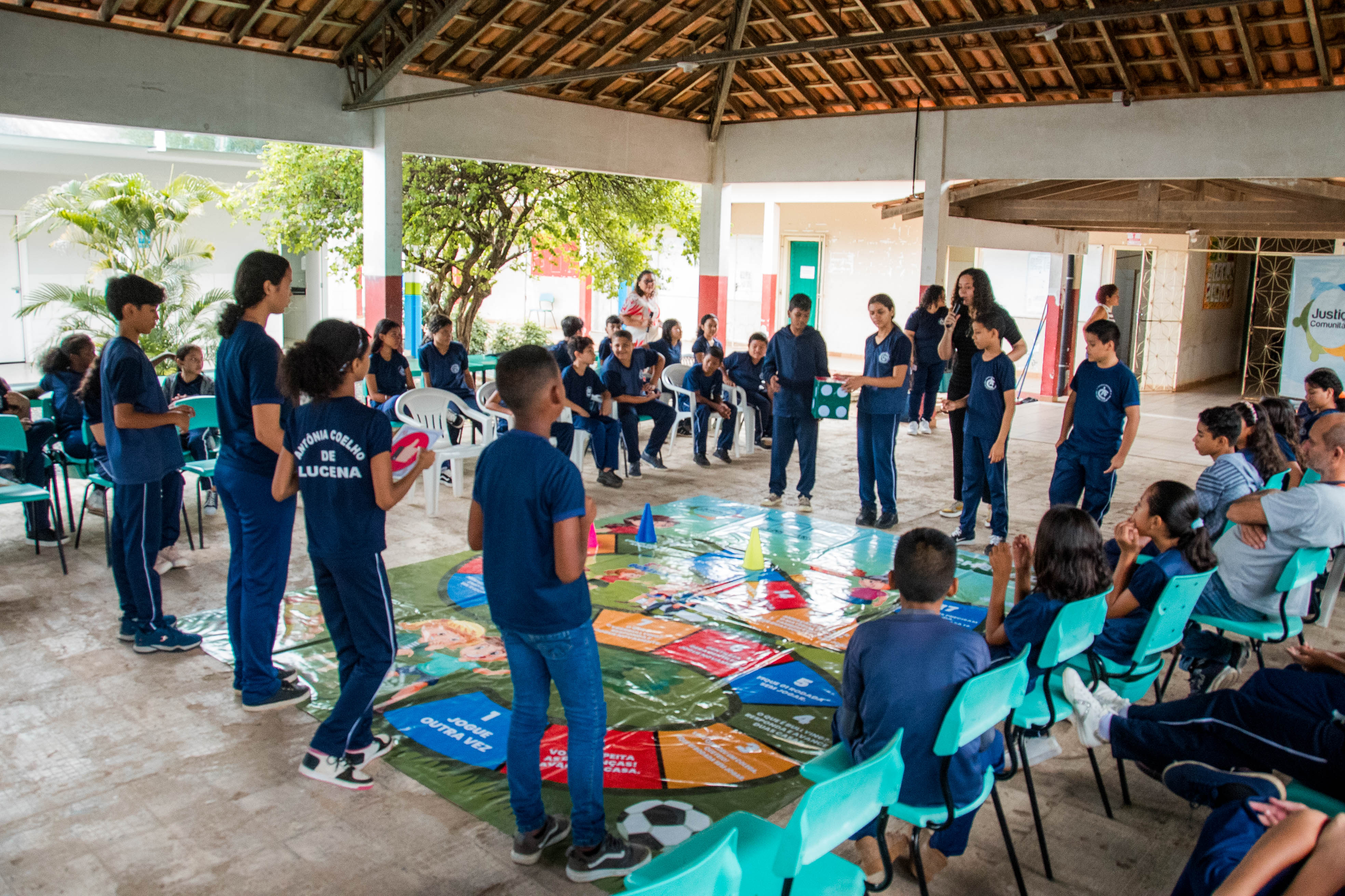 Alunos participam ativamente de uma dinâmica utilizando o tabuleiro gigante disposto no chão. Eles jogam um dado grande e interagem em grupo, enquanto outros assistem sentados ao redor. Alunos participam ativamente de uma dinâmica utilizando o tabuleiro gigante disposto no chão. Eles jogam um dado grande e interagem em grupo, enquanto outros assistem sentados ao redor.