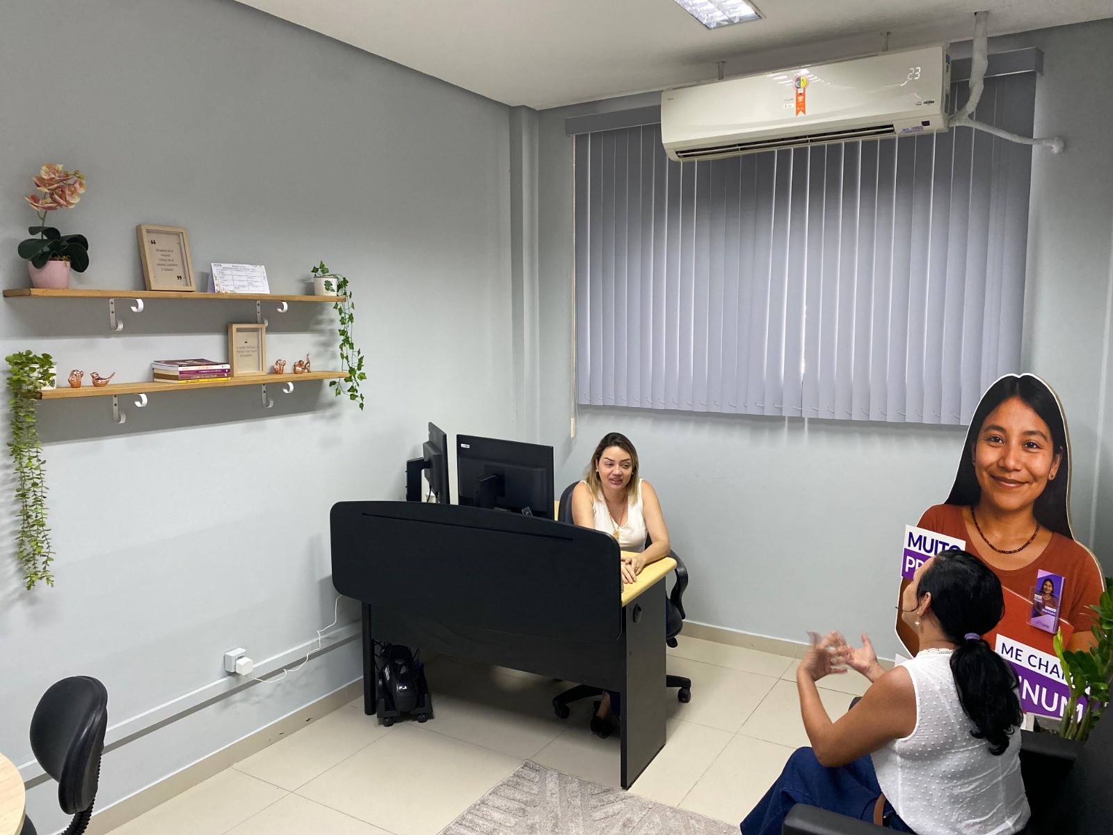 A imagem colorida mostra o interior de um escritório pequeno e bem iluminado. Há duas mulheres presentes conversando A imagem colorida mostra o interior de um escritório pequeno e bem iluminado. Há duas mulheres presentes conversando