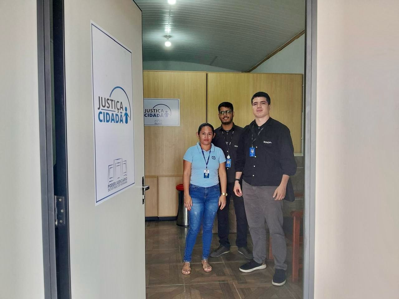 Imagem colorida mostrando uma porta aberta com dois servidores da equipe do TJRR pousando para fotografia ao lado de uma servidora do TJRR. Imagem colorida mostrando uma porta aberta com dois servidores da equipe do TJRR pousando para fotografia ao lado de uma servidora do TJRR.
