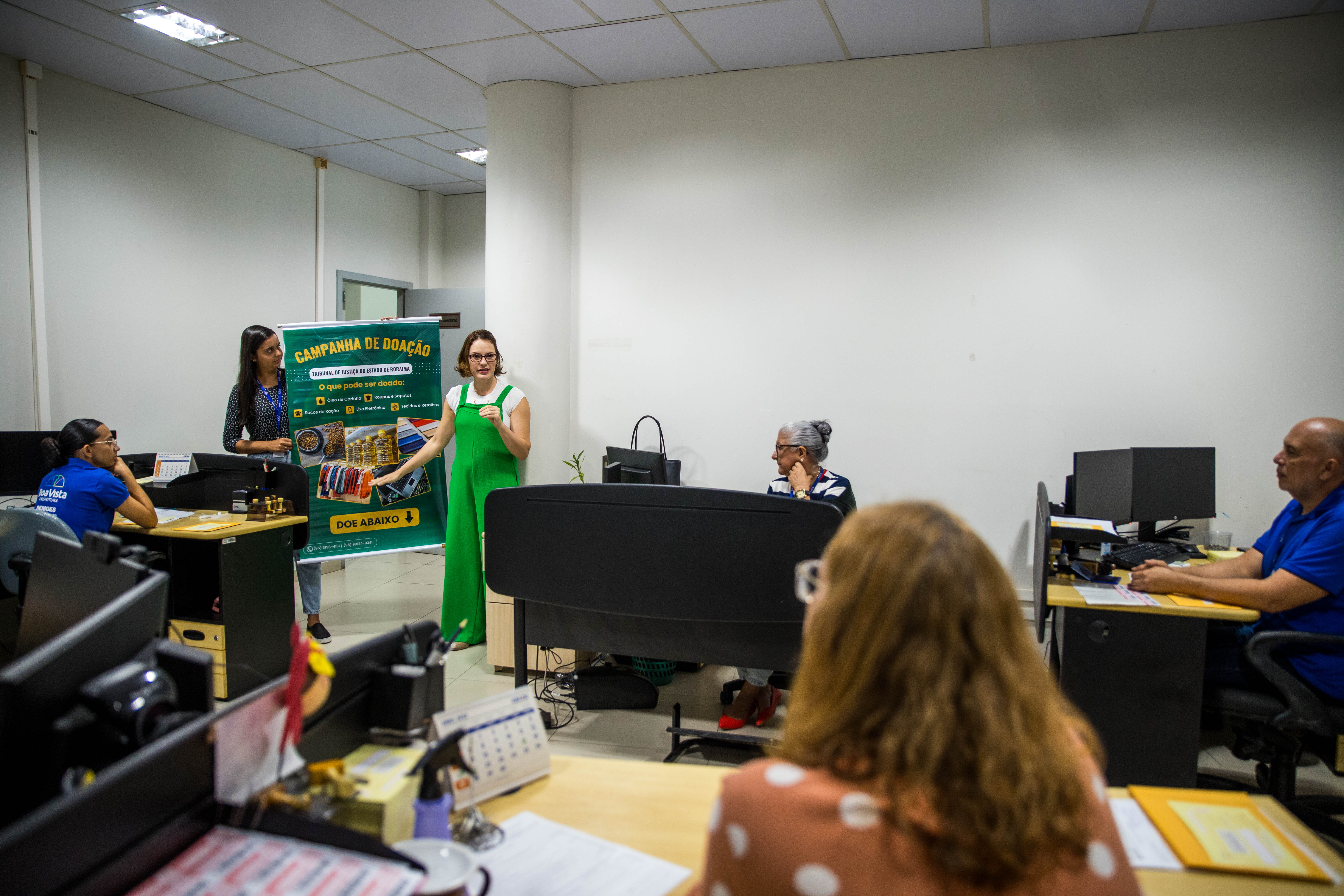 Foto colorida mostra uma mulher de macacão verde em um ambiente de escritório, no qual a mesma está apresentando uma campanha de doação para um grupo de funcionários. Ela está ao lado de um banner verde com informações sobre os itens que podem ser doados, como roupas, alimentos e livros. Algumas pessoas estão sentadas em suas mesas, ouvindo a apresentação, enquanto outra mulher segura o banner. Foto colorida mostra uma mulher de macacão verde em um ambiente de escritório, no qual a mesma está apresentando uma campanha de doação para um grupo de funcionários. Ela está ao lado de um banner verde com informações sobre os itens que podem ser doados, como roupas, alimentos e livros. Algumas pessoas estão sentadas em suas mesas, ouvindo a apresentação, enquanto outra mulher segura o banner.