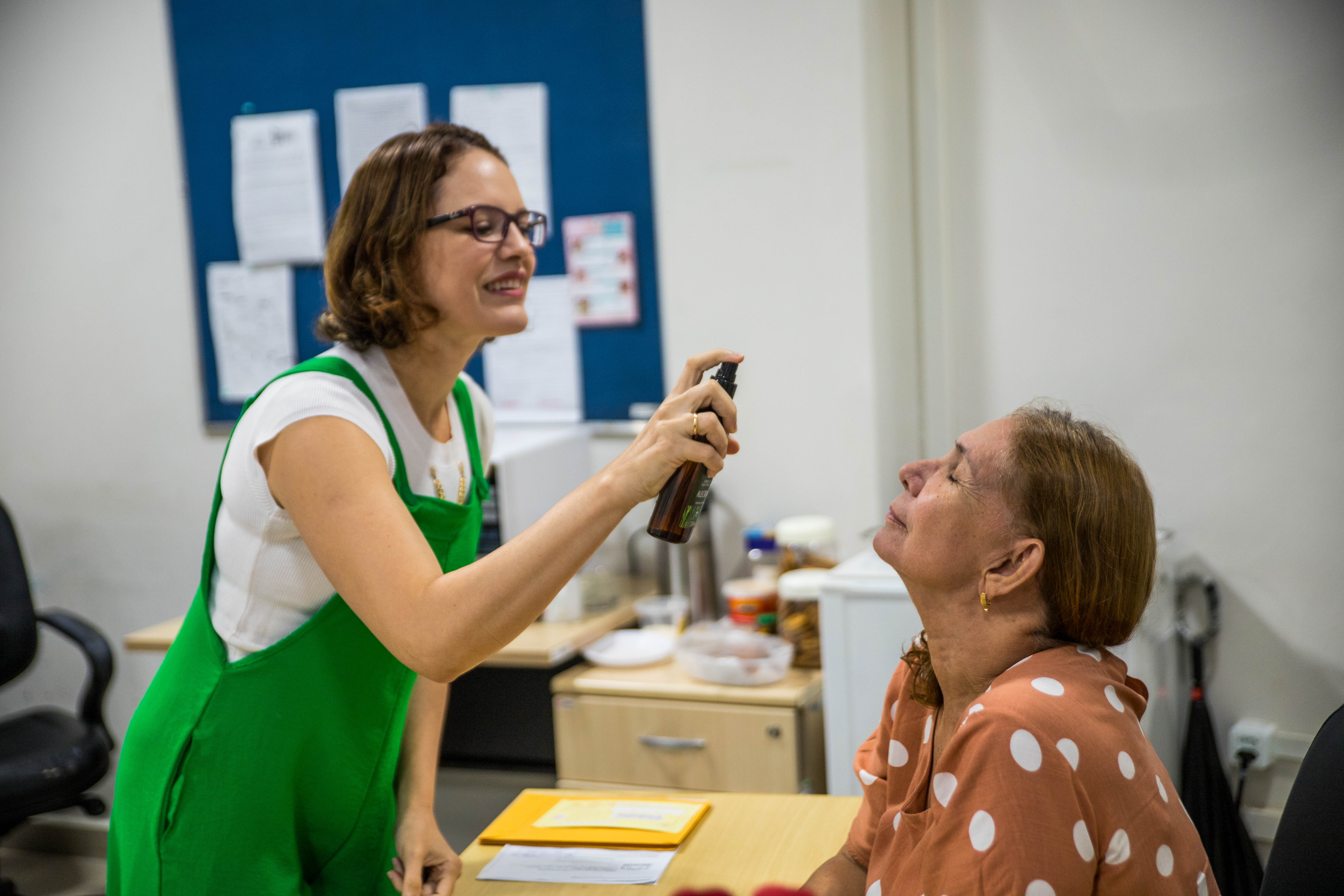 Foto colorida mostra uma mulher de macacão verde em um ambiente de escritório, no qual a mesma está aplicando um spray facial em outra mulher que está sentada à mesa, de olhos fechados e expressão relaxada. O clima da imagem transmite um momento de descontração no ambiente de trabalho. Foto colorida mostra uma mulher de macacão verde em um ambiente de escritório, no qual a mesma está aplicando um spray facial em outra mulher que está sentada à mesa, de olhos fechados e expressão relaxada. O clima da imagem transmite um momento de descontração no ambiente de trabalho.