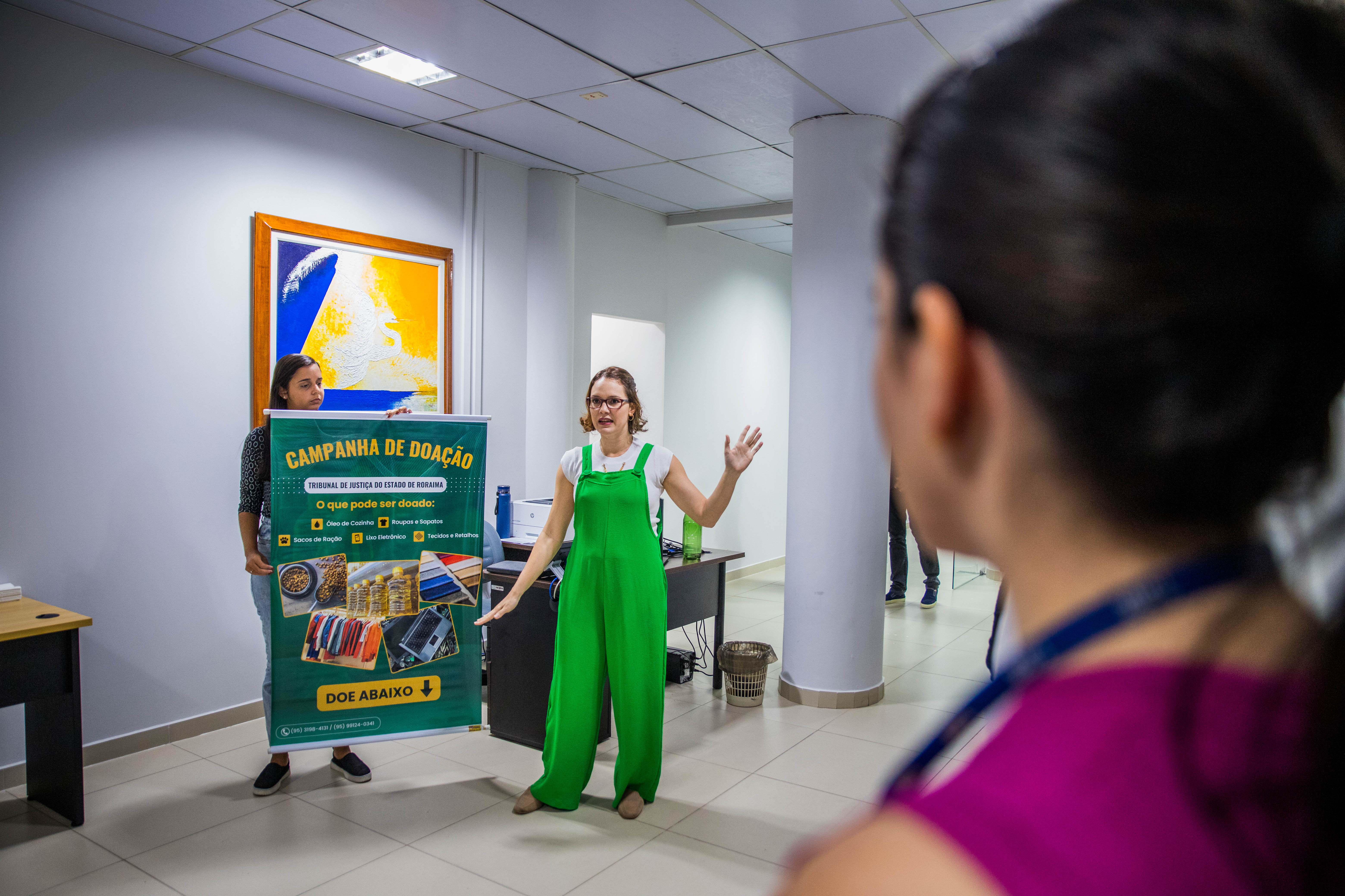 Foto colorida mostra a mesma mulher de macacão verde está explicando sobre a campanha de doação para um grupo de pessoas em um corredor do escritório. O banner da campanha está sendo segurado por outra mulher ao lado dela. Em primeiro plano, há uma pessoa de costas, ouvindo atentamente a explicação. Foto colorida mostra a mesma mulher de macacão verde está explicando sobre a campanha de doação para um grupo de pessoas em um corredor do escritório. O banner da campanha está sendo segurado por outra mulher ao lado dela. Em primeiro plano, há uma pessoa de costas, ouvindo atentamente a explicação.