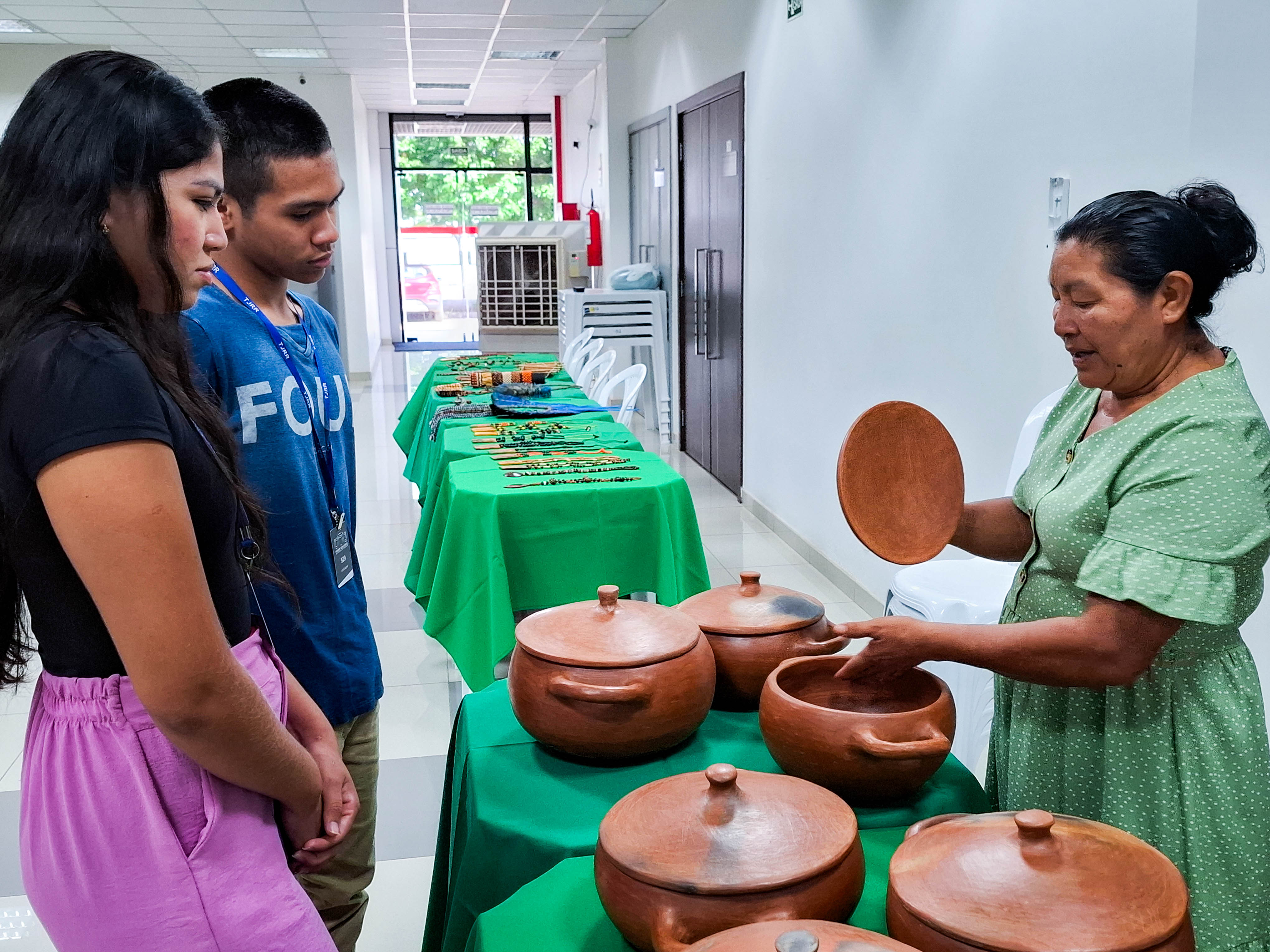 A imagem colorida mostra três indivíduos reunidos em torno de uma mesa coberta por uma toalha verde vibrante. Uma jovem está sentada à esquerda, enquanto um homem e uma mulher estão em pé à direita da mesa. A imagem colorida mostra três indivíduos reunidos em torno de uma mesa coberta por uma toalha verde vibrante. Uma jovem está sentada à esquerda, enquanto um homem e uma mulher estão em pé à direita da mesa.