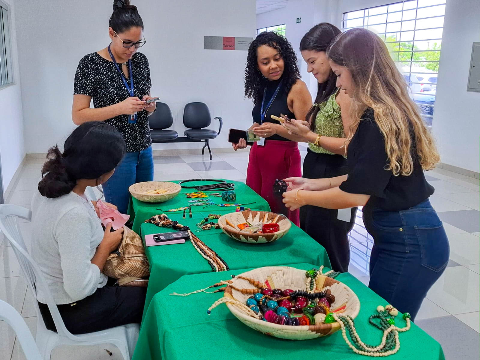 A imagem colorida mostra cinco mulheres reunidas em torno de uma mesa coberta por uma toalha de cor verde vibrante. Quatro mulheres estão em pé, observando e interagindo com os objetos dispostos sobre a mesa, enquanto uma quinta mulher está sentada à esquerda, de costas para a câmera, trabalhando em algo com as mãos. Na mesa, há diversos itens que parecem ser materiais para artesanato indígena. A imagem colorida mostra cinco mulheres reunidas em torno de uma mesa coberta por uma toalha de cor verde vibrante. Quatro mulheres estão em pé, observando e interagindo com os objetos dispostos sobre a mesa, enquanto uma quinta mulher está sentada à esquerda, de costas para a câmera, trabalhando em algo com as mãos. Na mesa, há diversos itens que parecem ser materiais para artesanato indígena.