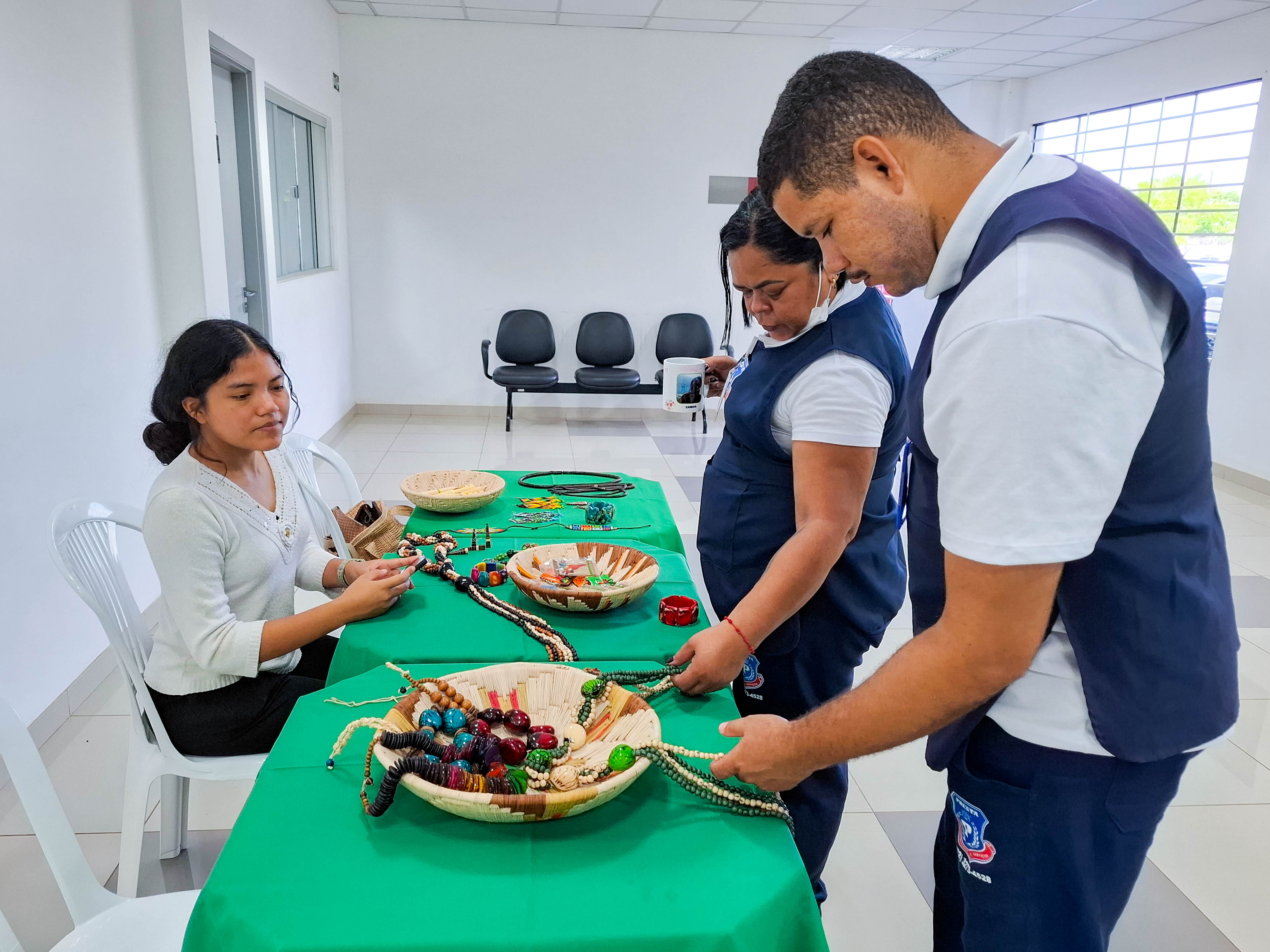 imagem colorida mostra três pessoas reunidas em torno de uma mesa coberta por uma toalha verde. Uma jovem está sentada à esquerda, enquanto um homem e uma mulher estão em pé à direita da mesa. imagem colorida mostra três pessoas reunidas em torno de uma mesa coberta por uma toalha verde. Uma jovem está sentada à esquerda, enquanto um homem e uma mulher estão em pé à direita da mesa.
