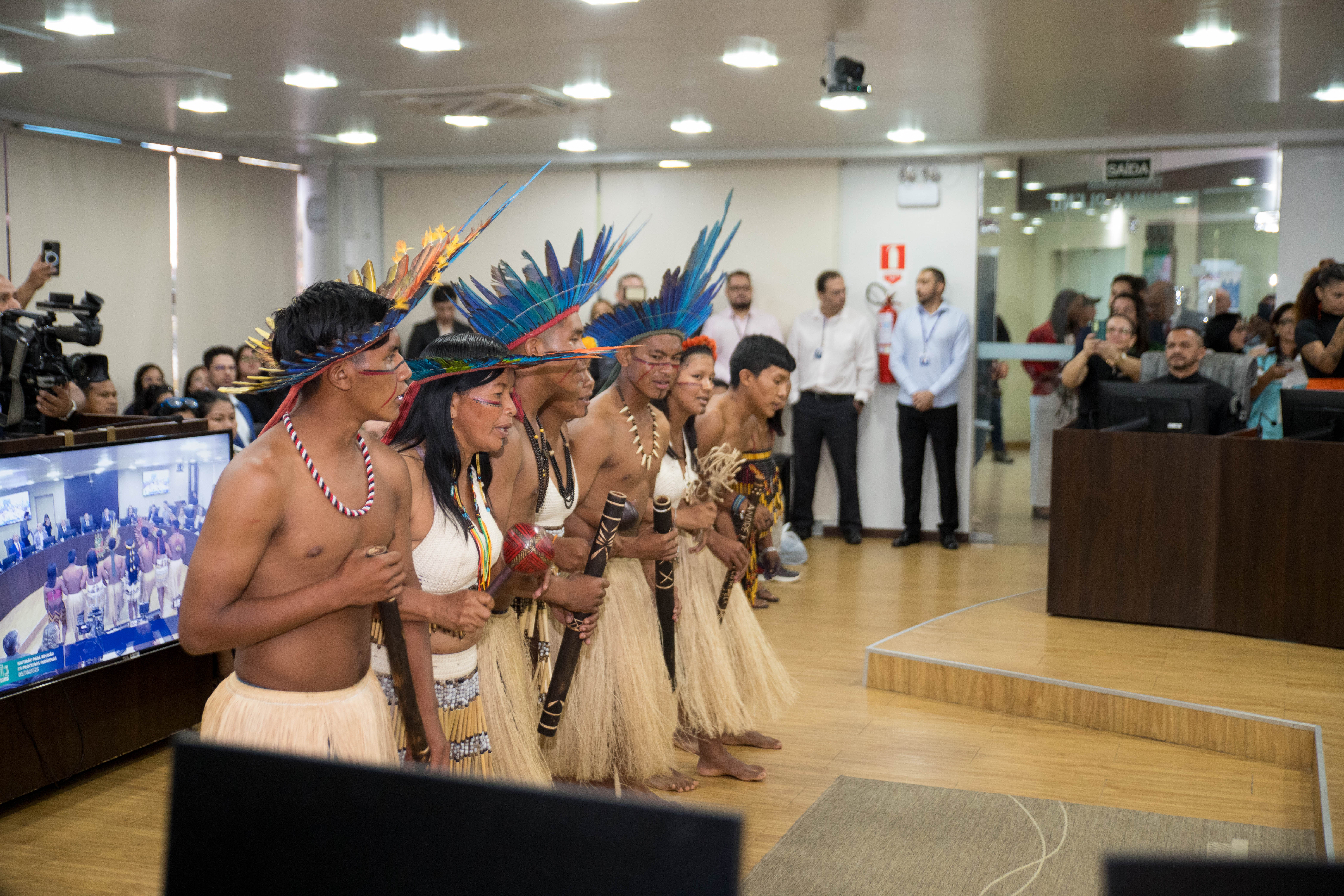A imagem retrata um grupo de pessoas indígenas na Sala de Sessões do Tribunal Pleno realizando uma apresentação cultural. A imagem retrata um grupo de pessoas indígenas na Sala de Sessões do Tribunal Pleno realizando uma apresentação cultural.